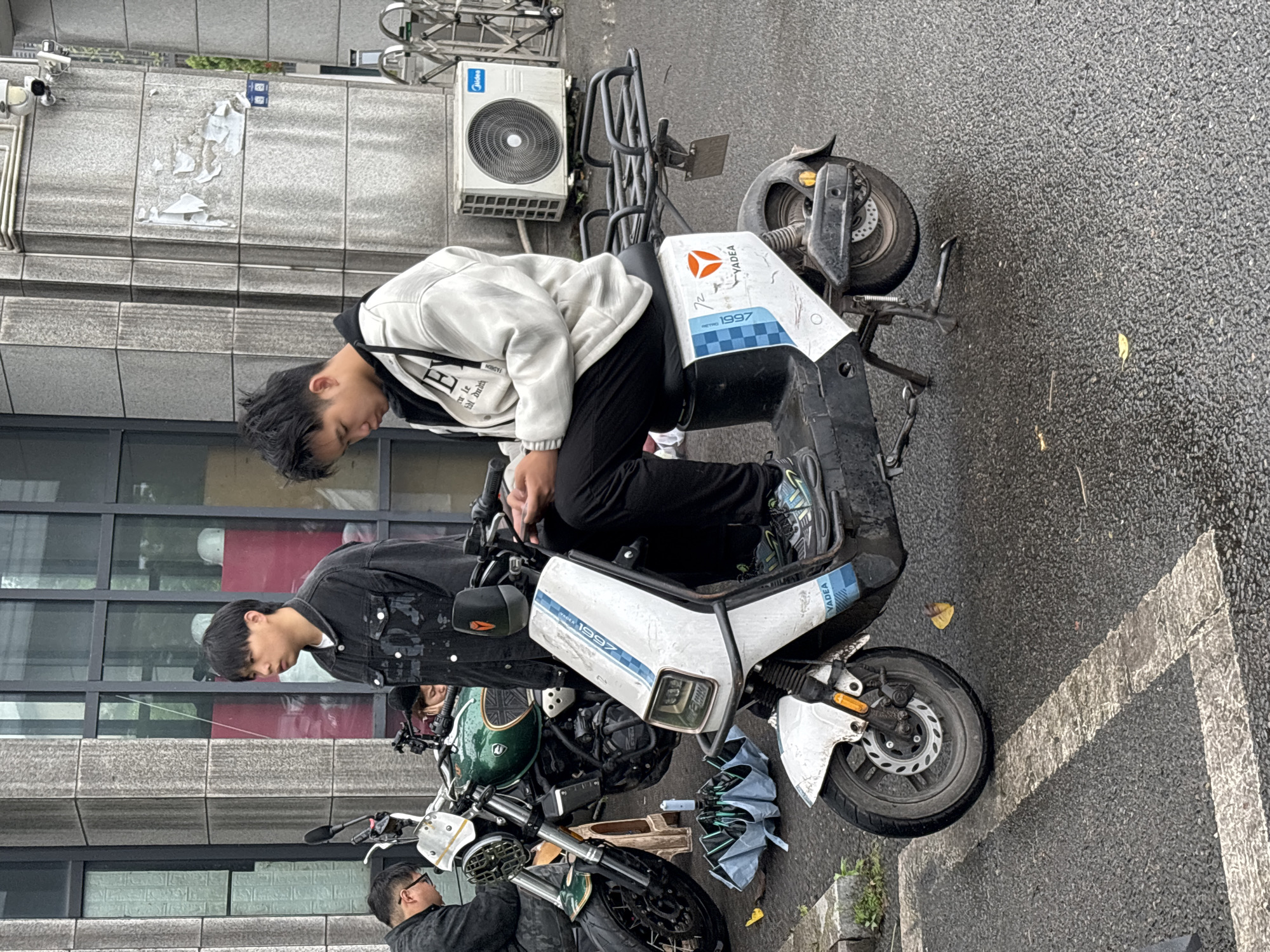 A medium shot, taken at a high angle, shows two Asian males interacting with motorized two-wheeled vehicles. The location is assumed to be Jiangjin, China, based on context.
A male wearing a white sweatshirt with black sleeves and black pants is sitting on a white Yadea brand scooter. He is looking down at the controls, seemingly inspecting or adjusting them.
Next to the scooter, another male wearing a black denim jacket, is standing next to a green and black motorcycle. He is also looking down, potentially making adjustments or repairs to the motorcycle.
A third male is mostly out of frame but appears to be interacting with the motorcycle.
The scooter and motorcycle are parked on a paved surface, likely a street or sidewalk. In the background, there is a building with glass windows and a wall with an air conditioning unit mounted on it. An umbrella is also present on the ground.