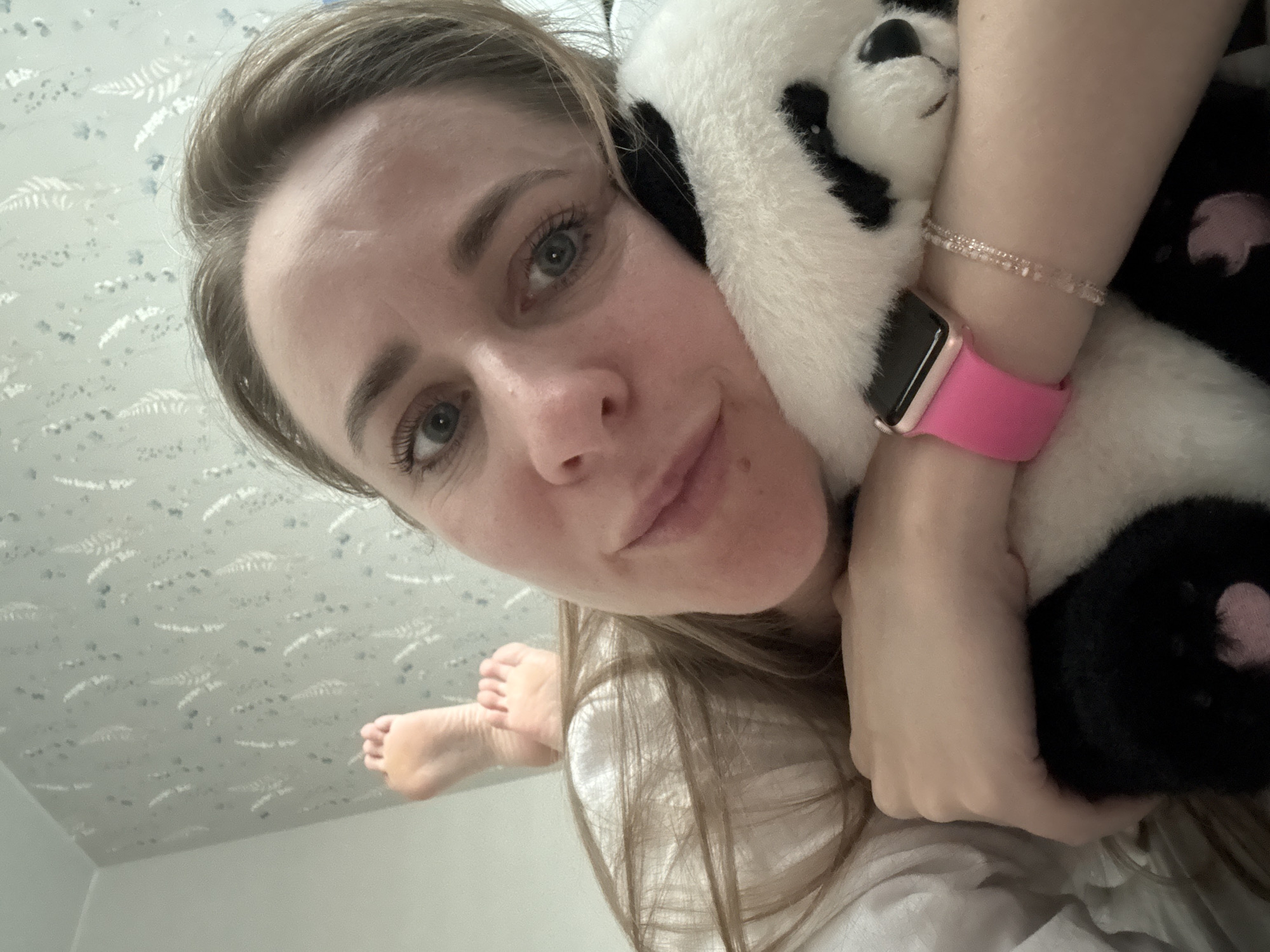 A light-skinned woman is holding a stuffed panda toy close to her face. She wears a pink Apple Watch on her left wrist, as well as a beaded bracelet. Part of a foot is visible in the background of the shot. The setting appears to be indoors in a room with patterned wallpaper. The location is unknown.