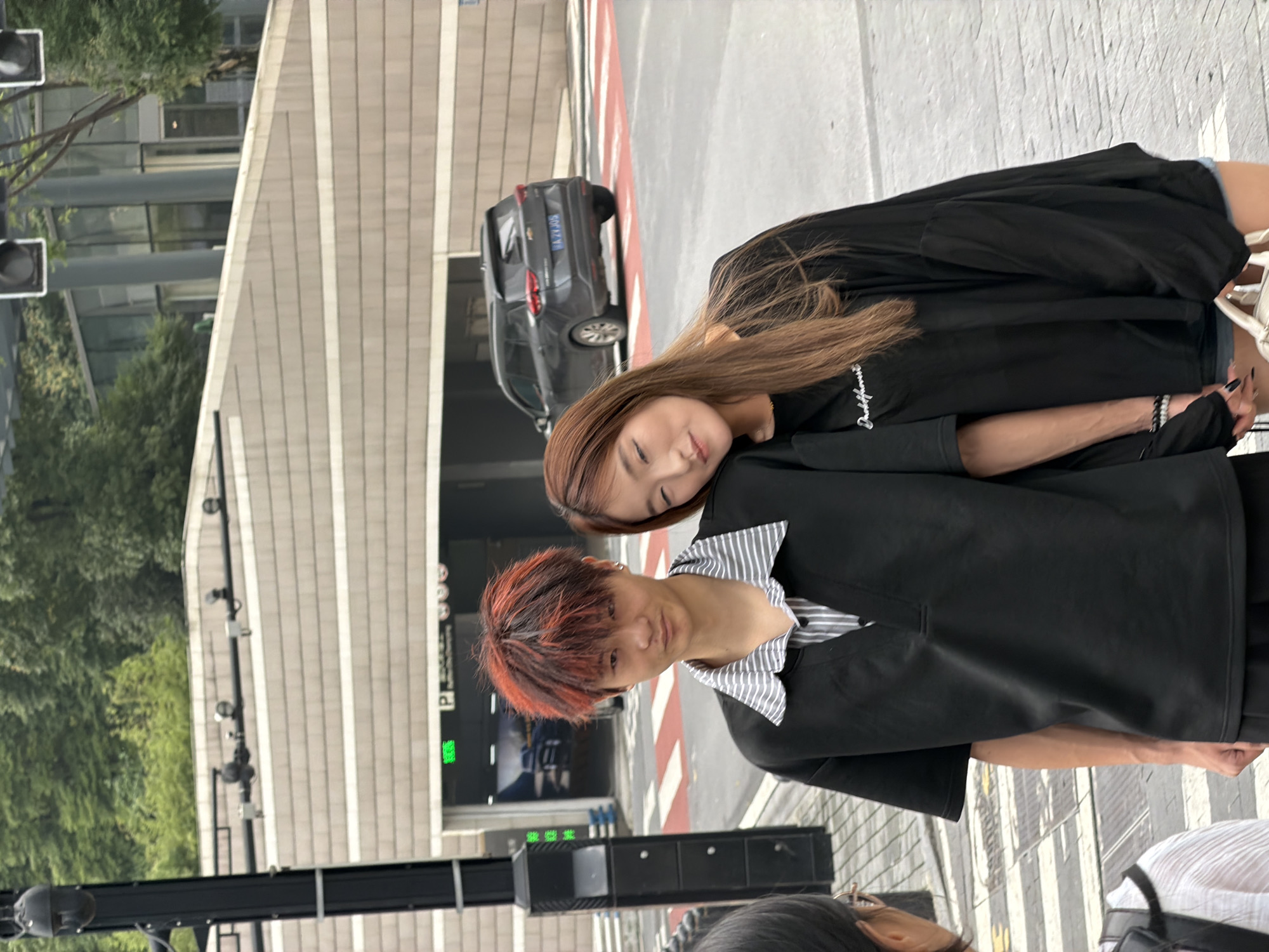 In Yuzhong District, China, a young Asian couple stands close together on a street. The man, with brightly dyed red and orange hair, wears a black collared shirt over a striped inner shirt. The woman, with long brown hair, leans her head on his shoulder, her eyes closed. They are both dressed casually. Behind them, a dark SUV is parked in front of a building entrance, indicated by a "P" sign and illuminated green lettering. The scene appears to be in an urban environment with a pedestrian crossing in the foreground and trees and buildings in the background.