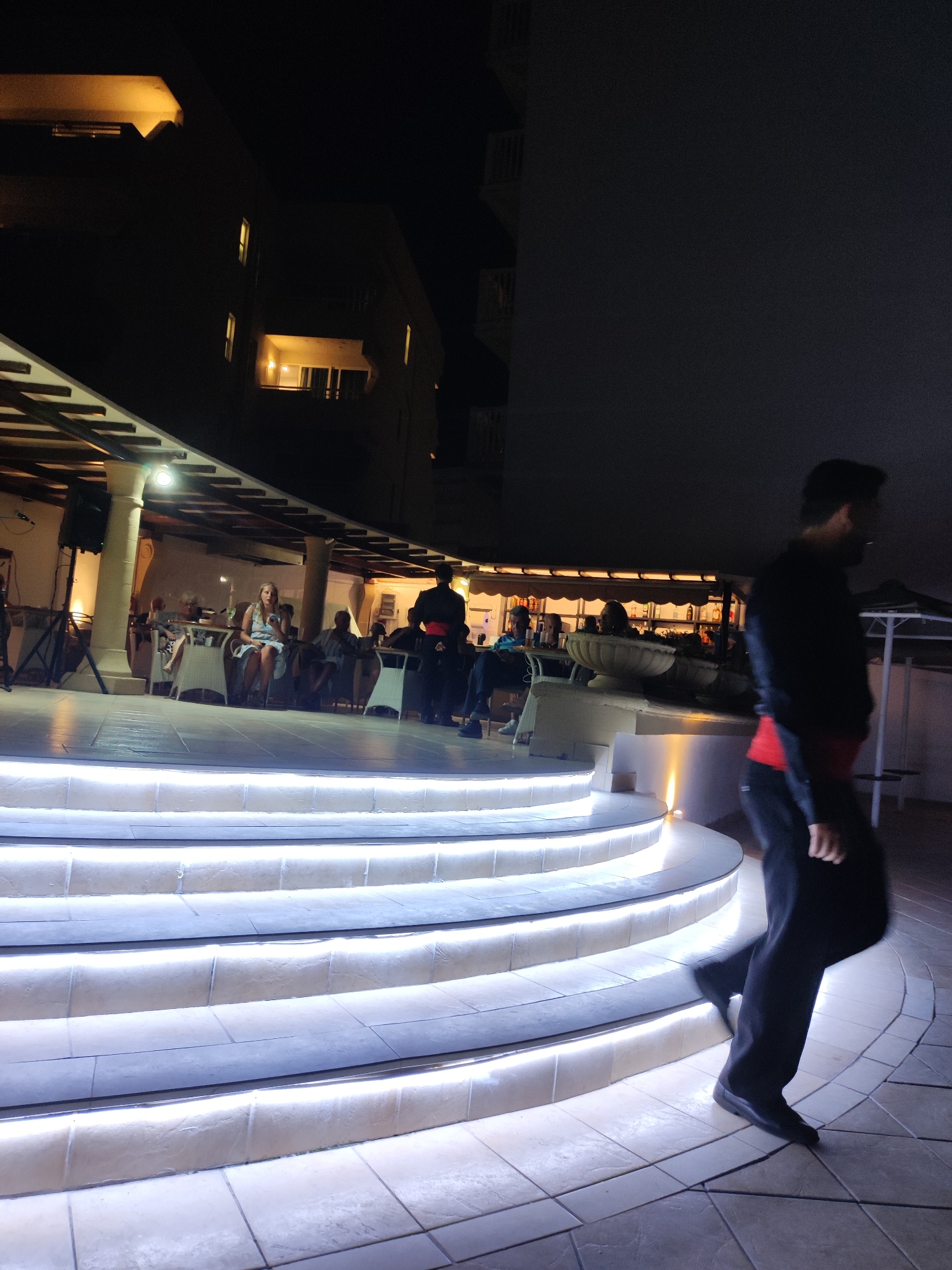 A man is walking down a curved staircase lit by white lights at night. He is wearing a black suit with a red sash. In the background, there is a restaurant with people seated at tables and a woman speaking into a microphone. The restaurant appears to be outdoors, with a covered patio. The scene takes place in the Municipal Unit of Rhodes, Greece.