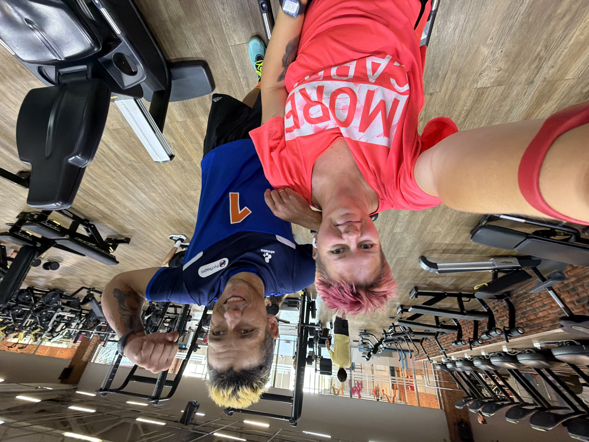 The image depicts an indoor gym with wooden flooring. In the foreground, a woman with short, pink hair is seated in a wheelchair, wearing a bright pink t-shirt with white text. She is holding a red resistance band around her arm and appears to be taking a selfie. Next to her, a man with spiked blonde and brown hair, wearing a blue athletic shirt and black shorts, is flexing his bicep and smiling. Behind them, various gym equipment is visible, including treadmills, weight machines, and benches, arranged in rows. In the background, other individuals can be seen exercising or walking around the gym. The overall impression is one of a fitness facility. The text on the man's shirt includes Cyrillic characters and the word "PROSPORTS". Based on contextual clues, the location is identified as the "SPARTA" gym in Chaikovsky, Russia.