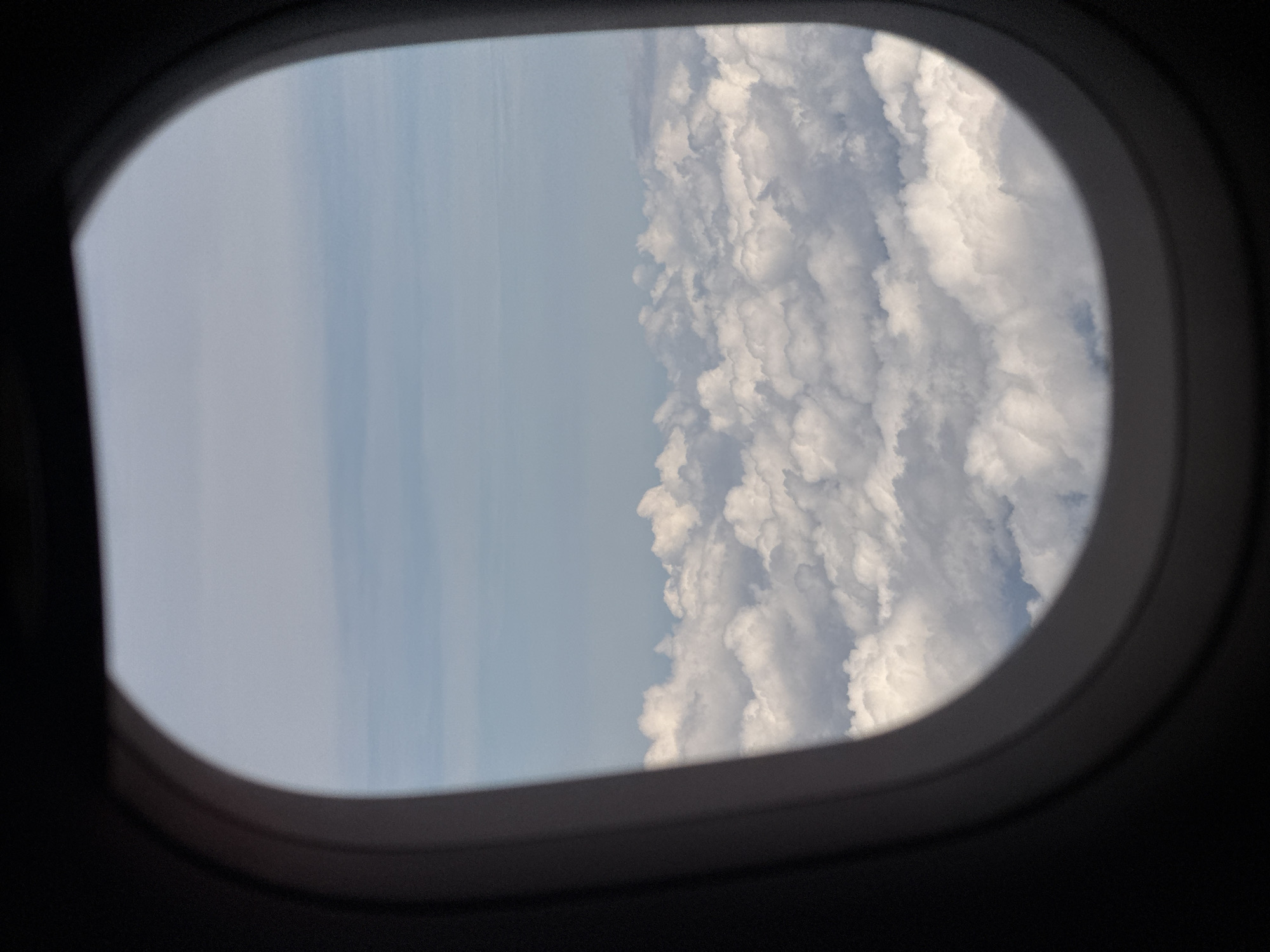 A view from an airplane window shows a dense layer of white, fluffy clouds stretching across the horizon, with patches of blue sky visible above. The window frame, a dark grey oval, partially obstructs the view. The image is taken from an aerial perspective, suggesting the airplane is flying above the cloud layer. There are no discernible people, objects, or actions visible within the image. The location in Ban Na Thao, Thailand, is not evident from the visual information presented.
