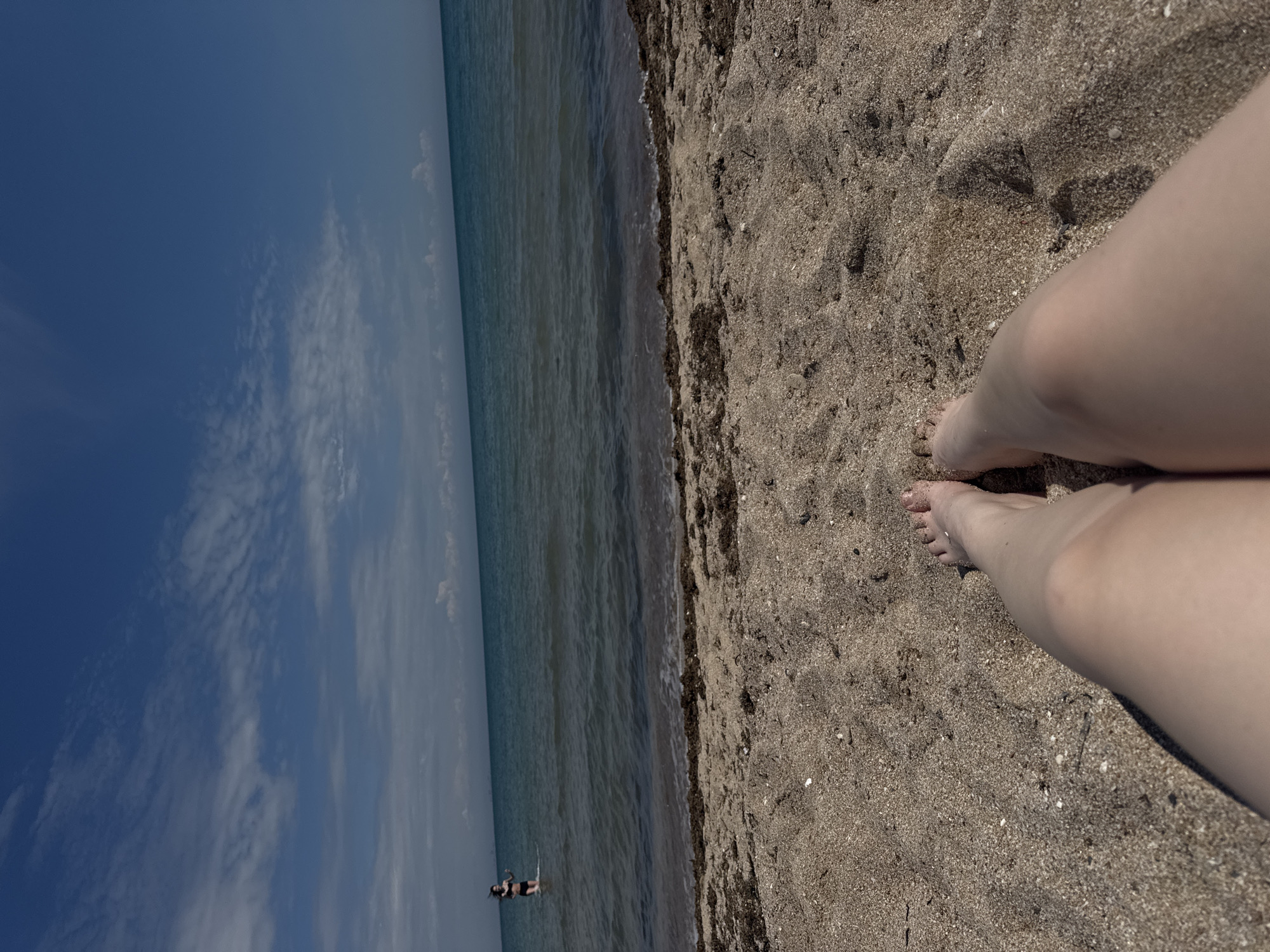 The image shows a view of the Black Sea from a beach in Yevpatoriya, Ukraine. The perspective is from someone standing on the sandy beach, with their legs and feet visible in the lower right. The sand is coarse and speckled with shells and dark debris. The sea stretches along the left side, with calm water meeting the sandy shore. In the water, there appear to be two figures, possibly people swimming or standing. The sky is a clear blue with scattered white clouds. The photo is rotated counter-clockwise, causing the horizon to appear vertical.