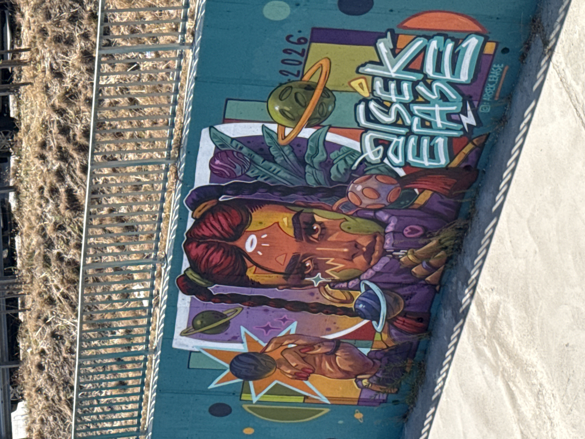 The image depicts a colorful mural painted on a concrete wall in Sofia, Bulgaria. The mural features a stylized portrait of a person with multiple eyes and elaborate braided hair, surrounded by celestial elements like planets and stars. The color palette is vibrant, with prominent use of teal, orange, purple, and yellow. In the background, there is a metal railing and dry vegetation. The mural also includes the text "2026" and a signature that appears to be "@ARSEK.ERASE". The lower part of the image shows a concrete pathway casting shadows from the railing.