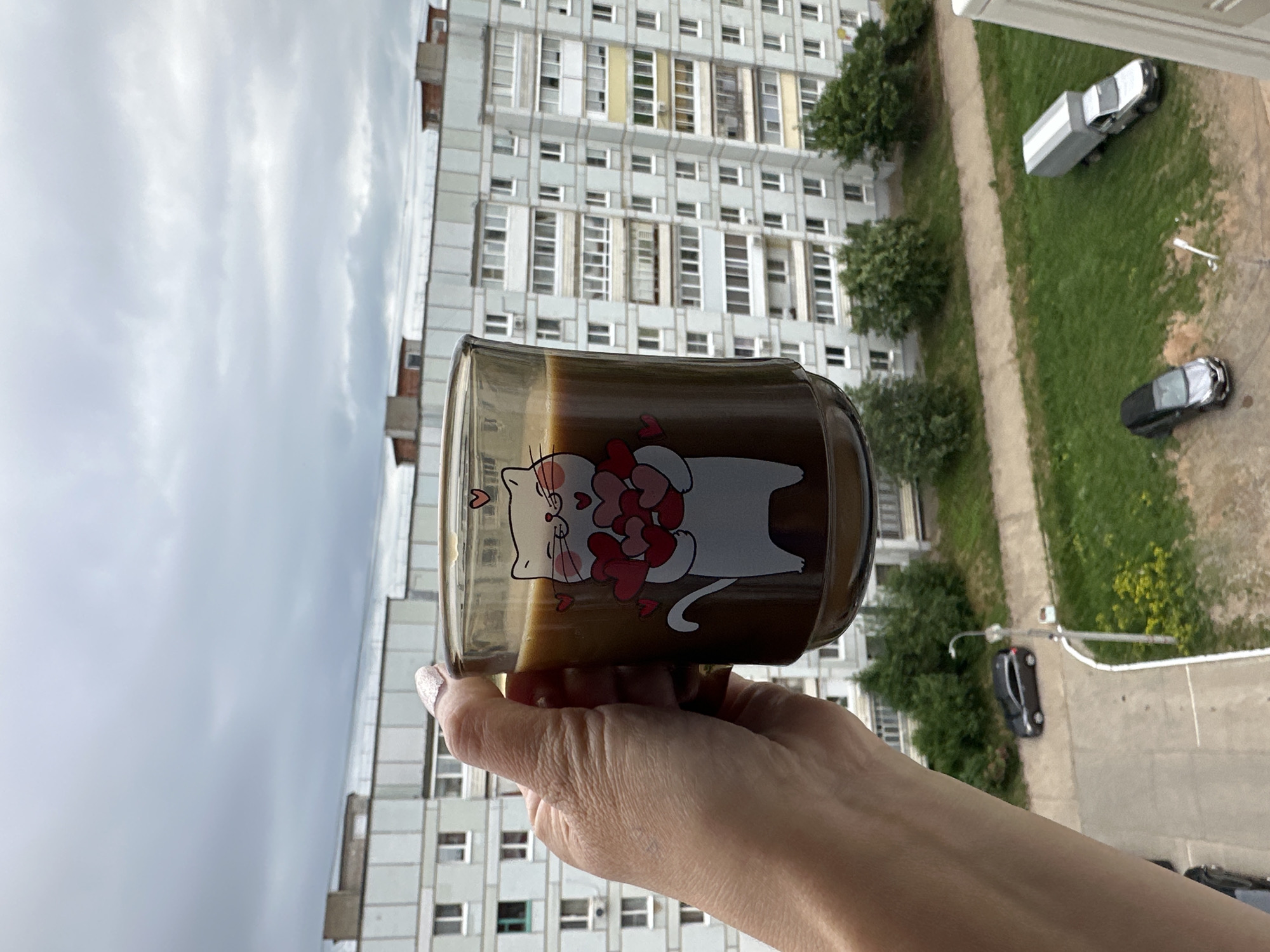 A hand holds a clear glass mug filled with a light brown beverage. The mug features a white cartoon cat design with pink hearts. The background depicts a tall, multi-story apartment building with numerous windows, typical of Soviet-era architecture. Green trees and grass are visible between the building and the street. On the street, there are several cars, including a black hatchback and a silver truck. The sky is overcast. The scene appears to be in a residential area of Chaikovsky, Russia.