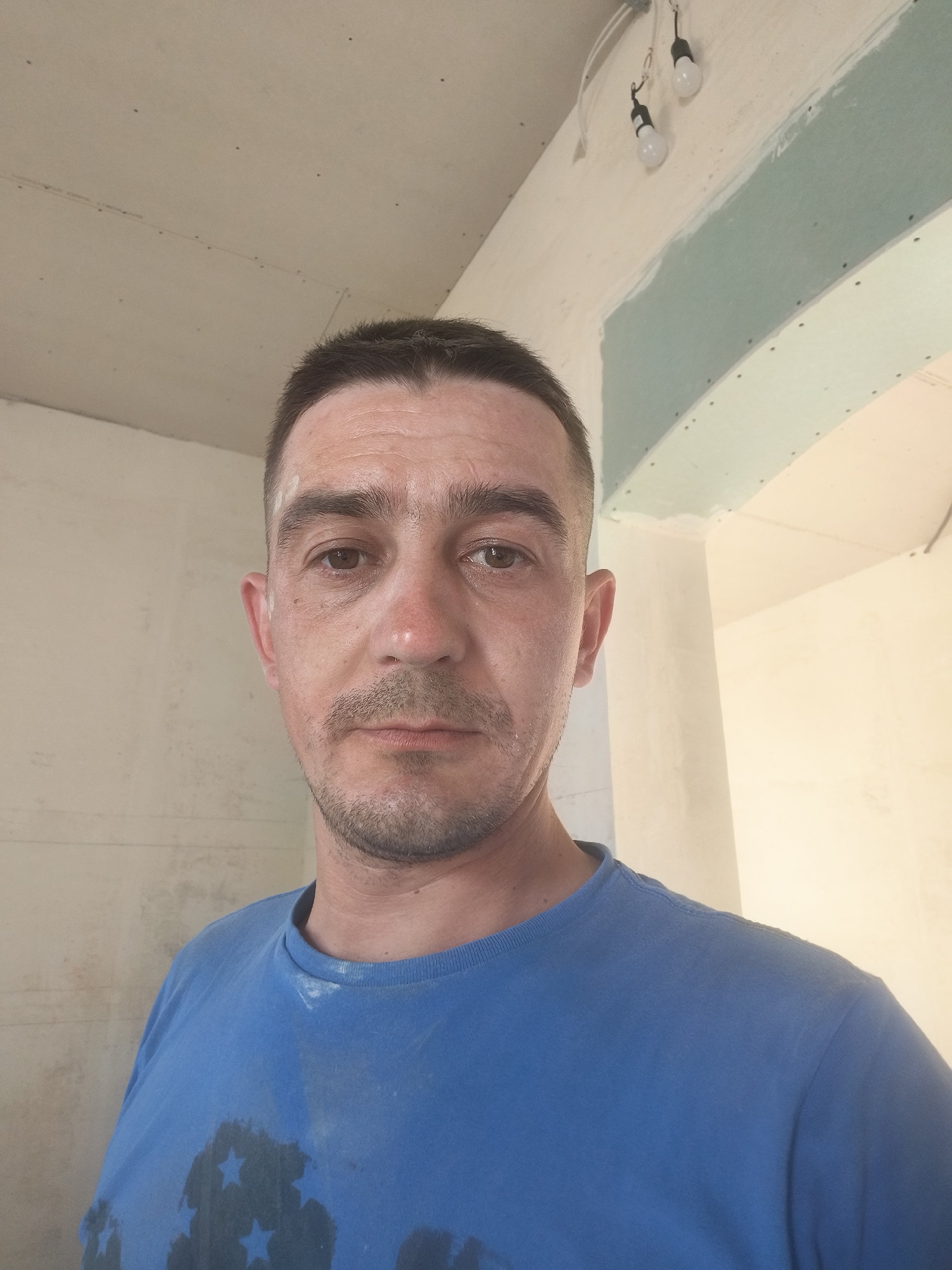 A close-up, selfie-style photograph shows a fair-skinned man with short, dark hair, a short beard, and light brown eyes. He is wearing a blue t-shirt with a faded dark star pattern. The man appears to be in an unfinished interior space, likely a building under construction or renovation. The walls and ceiling are bare, with visible drywall and some light fixtures hanging from the ceiling. The overall impression is one of a candid, informal moment captured during what appears to be manual labor. There are no discernible landmarks or contextual clues that would place this image specifically in Grinăuți, Moldova.