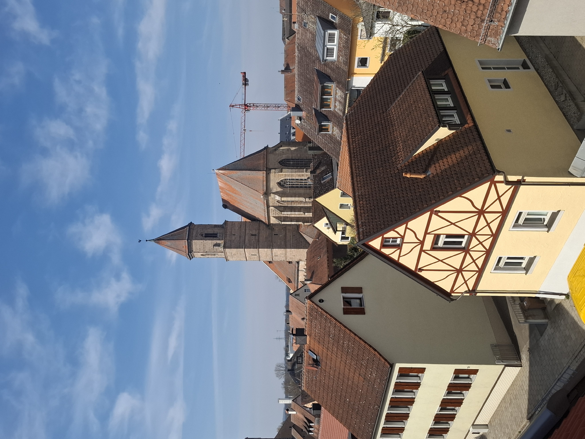 An elevated, clear-day view depicts a dense collection of buildings in Gunzenhausen, Germany, under a bright blue sky with scattered white clouds. The scene is devoid of visible people or active vehicles.
In the foreground, a light beige residential building with a reddish-brown tiled gable roof occupies the left side. It features multiple rectangular windows with closed dark brown wooden shutters on its two visible stories, and a white brick-patterned base. Adjacent to it, a light yellow half-timbered house with dark brown wooden beams arranged in cross and diagonal patterns stands prominently on the right, also with a reddish-brown tiled gable roof. This house displays white-framed windows, some with dark brown shutters, and a dark brown downspout. A paved sidewalk runs between these buildings. A small segment of a bright yellow tarp or structure is partially visible at the bottom center.
The midground is filled with numerous other residential structures, primarily featuring reddish-brown tiled roofs, some with dormer windows, chimneys, and satellite dishes.
Dominating the background and center is the St. Michael's Church, a large stone structure with a tall, multi-tiered square bell tower culminating in an octagonal, red-tiled spire topped by a finial, likely a weathervane. The church's nave section, constructed of stone, features prominent arched windows and a steeply pitched reddish-brown tiled roof. To the right of the church, a large red lattice-boom construction crane extends into the sky. The distant horizon reveals faint outlines of trees and potential industrial elements. Shadows cast by the buildings indicate strong sunlight from the left.