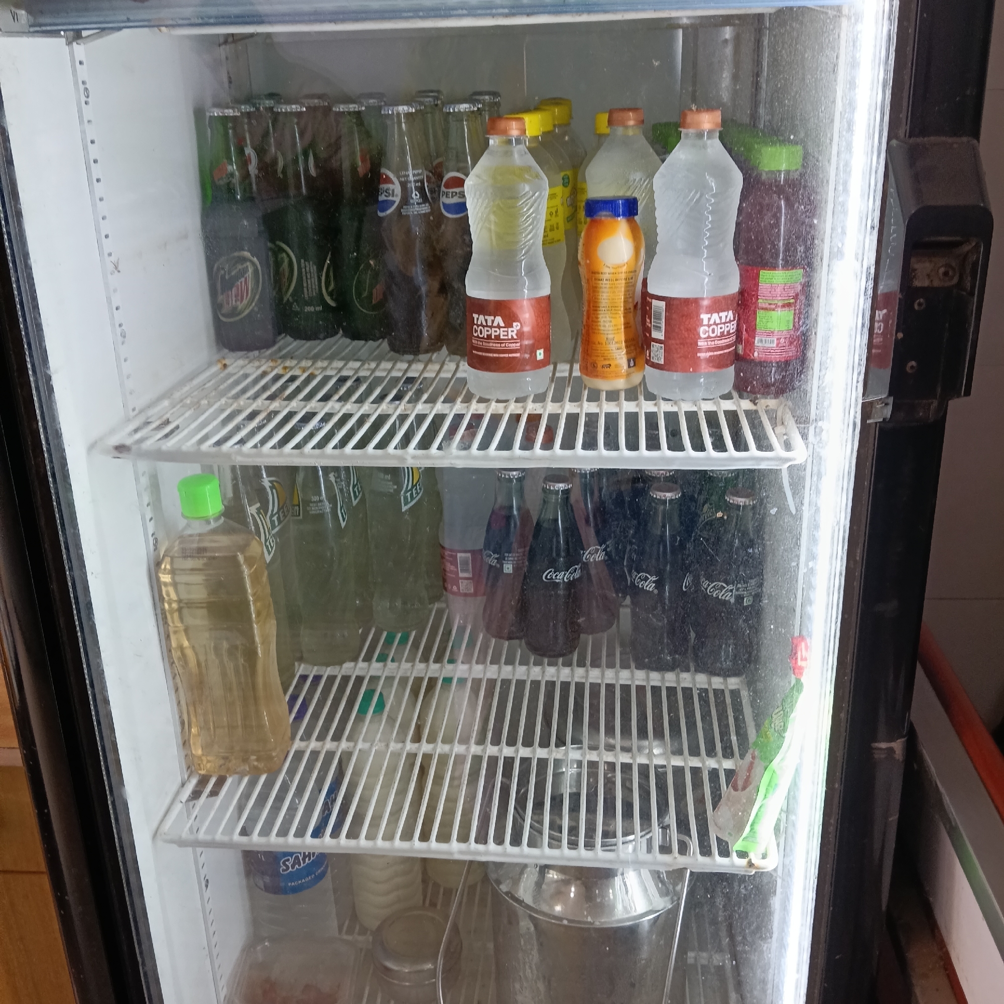 An open refrigerator is stocked with assorted beverages. On the top shelf are glass bottles of Mountain Dew and Pepsi, alongside plastic bottles labeled "TATA Copper" and other assorted juices. The middle shelf contains more plastic bottles filled with liquid and glass bottles of Coca-Cola. The bottom shelf has additional beverages and other food containers, including a stainless steel pot. The refrigerator is located in Oxel, India.