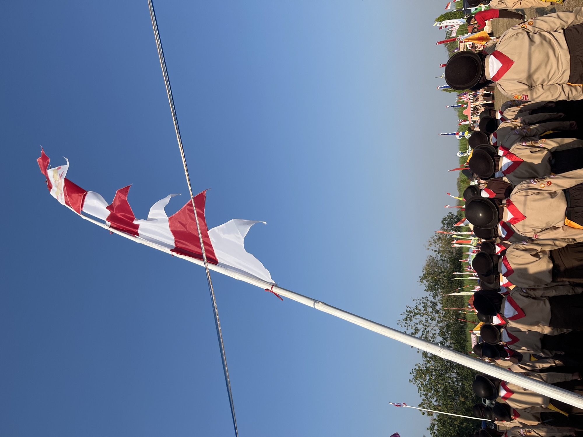 The image shows a group of scouts in uniform, standing in formation outdoors. They are wearing tan shirts with red and white neckerchiefs, and dark berets. A tall white flagpole with red and white flags is prominently displayed in the foreground, angled against a clear blue sky. In the background, a line of trees and other flags can be seen. The scene appears to be a formal gathering or event. The location is identified as Gembong, Indonesia, which is a sub-district in Pati Regency, Central Java Province.
