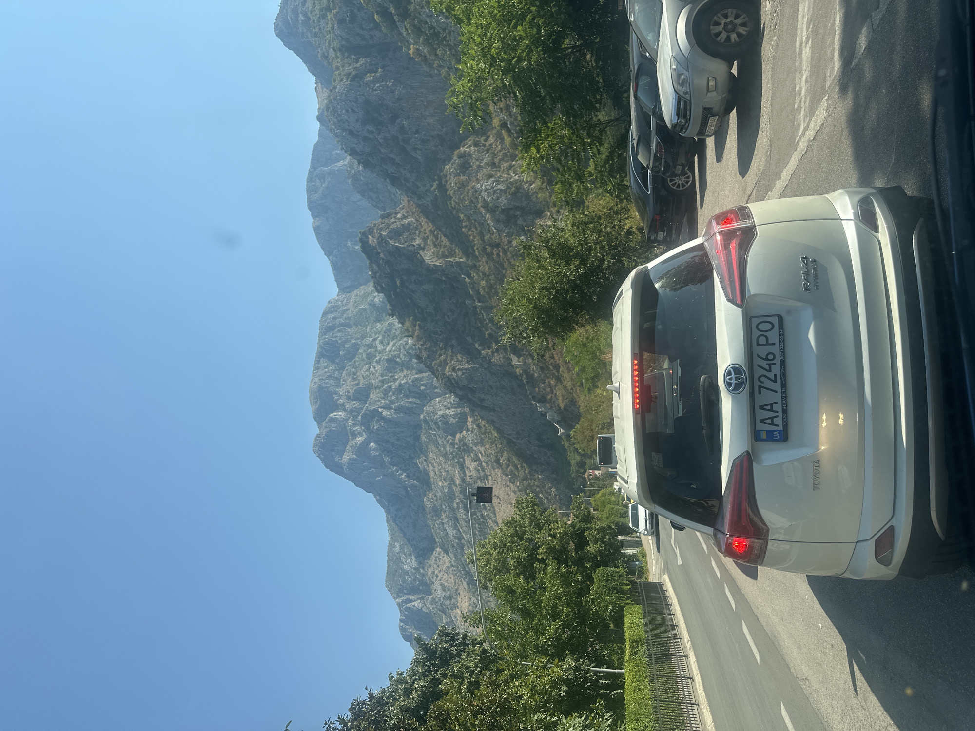 The image captures a view from a vehicle in traffic on a sunny day, likely in Škaljari, Montenegro. The foreground is dominated by the rear of a white Toyota RAV4 Hybrid, showing its license plate which reads "AA 7246 PO" with Ukrainian plates. To the left, a road curves away, bordered by a black metal fence and green hedges. A white car is visible further up the road. To the right, several other cars are parked or in traffic. In the background, a majestic mountain range with rocky, arid slopes rises under a clear blue sky. Lush green trees line the sides of the road, creating a contrast with the stark beauty of the mountains. A traffic light with a pedestrian signal is visible above the road. The overall impression is one of a scenic mountain town experiencing moderate traffic on a clear, warm day.