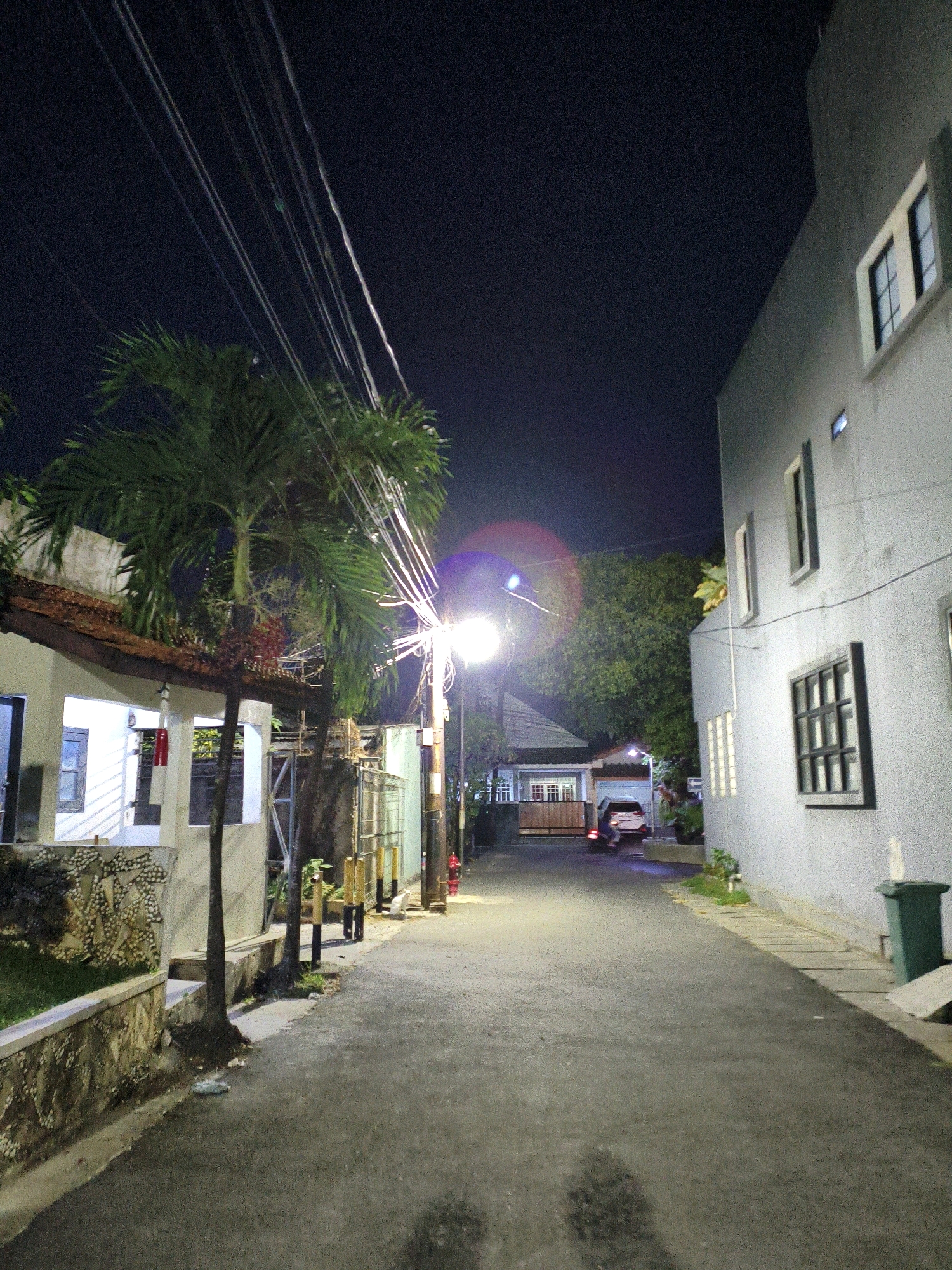 The image depicts a street scene at night in Bekasi, Indonesia. A narrow asphalt road runs through the middle of the frame, flanked by buildings and vegetation. On the left, a single-story building with a white facade and a tiled roof is visible. It features several windows, one with a dark frame. Adjacent to this building is a stone retaining wall with grassy areas and a small palm tree.
To the right, a multi-story building with a light gray textured facade dominates the foreground. This building has several evenly spaced rectangular windows, some with dark frames and others with lighter frames. A green trash bin is situated near the right edge of this building.
The road itself is illuminated by a street light positioned mid-frame on the left, casting a bright glow and creating a lens flare in the upper right quadrant of the sky. Several utility wires crisscross the dark night sky above the palm tree and the street. In the distance, at the end of the road, a white car with its taillights illuminated is parked or slowly moving. Further back, another building with multiple windows is visible. The overall impression is one of a residential street in an urban or suburban area, lit by artificial light against the dark night sky.