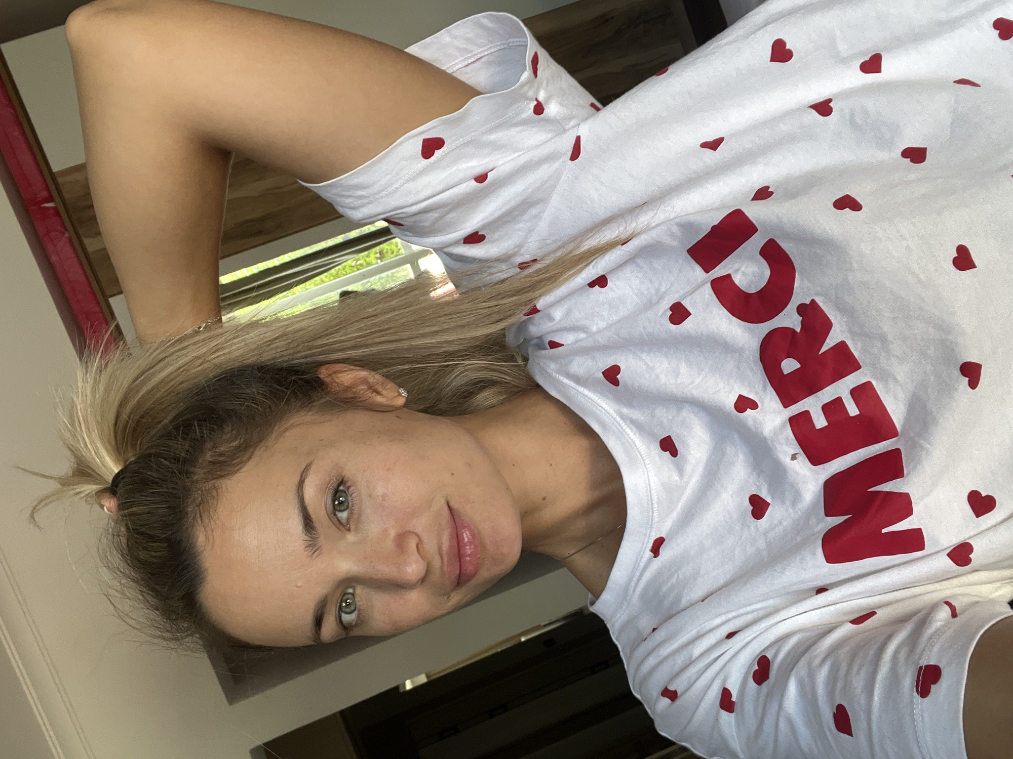A blonde woman is taking a selfie, wearing a white t-shirt with small red hearts and the word "MERCI" in red letters. Her hair is pulled up in a ponytail, and her left arm is bent behind her head. She has light skin, green eyes, and light freckles across her nose and cheeks. Her lips are slightly parted, and she is looking directly at the camera with a subtle smile. The background appears to be an indoor setting, possibly a bedroom, with a hint of a window showing greenery and sunlight. The image does not contain information indicating its location in Sharm El Sheikh, Egypt.