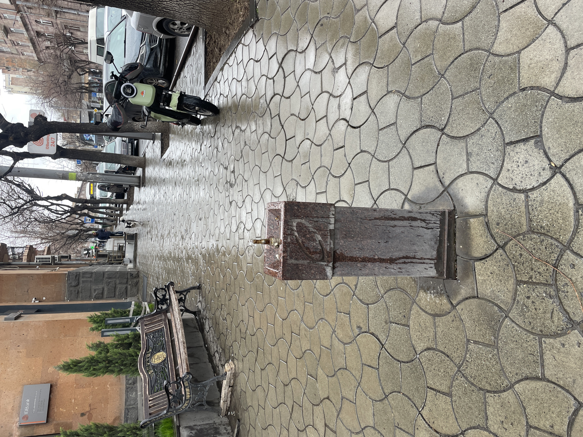 The image presents a wet urban sidewalk in Yerevan, Armenia. In the immediate foreground, a reddish-brown granite column, approximately waist-height, serves as a drinking fountain (*pulpyulak*), featuring a brass faucet at its top and dark water stains streaking down its front. The sidewalk is paved with light grey interlocking concrete pavers forming a scalloped pattern, thoroughly wet and reflective. To the left, a dark brown wooden park bench with ornate black metal scrollwork arms and legs, and a central circular emblem, is positioned next to a planter containing three conical evergreen trees. Behind this, a building with an orange-brown stucco façade displays a dark grey "IDBank" sign with white text, including Armenian script. To the right, a light green electric scooter with a circular LED headlight and "4G" branding is parked on its kickstand on the sidewalk. Multiple bare-branched deciduous trees line the street, behind which numerous cars are parked parallel to the curb. Further down the sidewalk, a male individual in a dark jacket is visible walking away from the camera, accompanied by an unleashed black and white dog. The sky is overcast, and the overall environment is typical of an urban street after rain.
