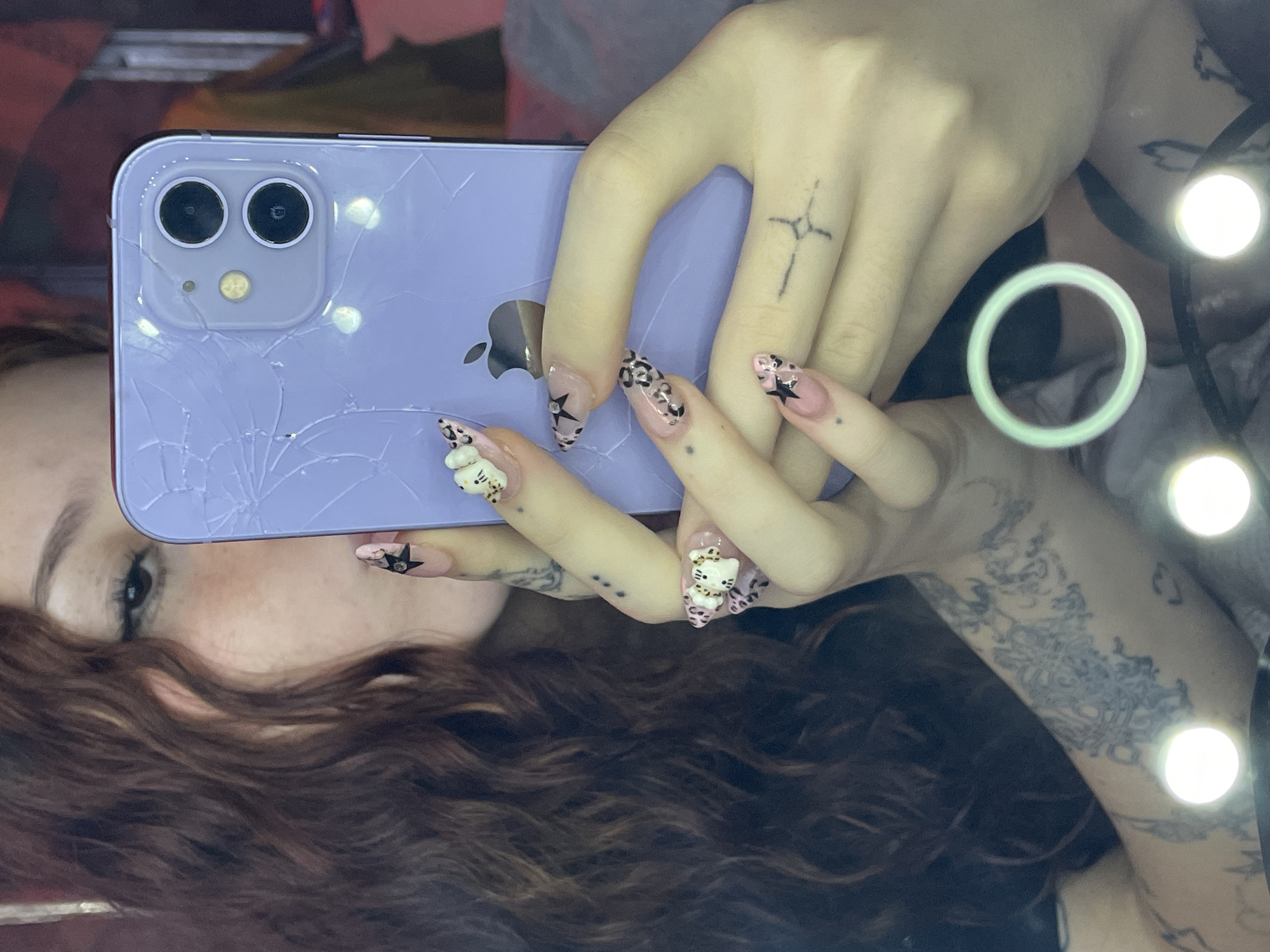 A mirror selfie captures a person holding a purple iPhone with a severely cracked back, particularly around the dual camera lenses and the lower section. The individual's hands are prominently featured, adorned with elaborate nail art. Some nails display black stars on a light pink base, others feature black and pink leopard print, and at least two nails have white 3D Hello Kitty charms.
The person's right hand (viewer's left) has a minimalist cross tattoo on the middle finger. The left hand (viewer's right) shows three small dot tattoos on the ring finger. A larger, intricate tattoo is visible on the person's left forearm/wrist area. Only a portion of the person's face is visible, including an eye with eyeliner and long, wavy dark brown hair cascading into the foreground.
The background is a reflection, showing multiple bright, circular light sources, including a distinct light green ring light. The Apple logo is visible on the back of the iPhone. The lighting suggests an indoor setting, and the selfie is being taken in front of a reflective surface. The context provided indicates this scene is in Buenos Aires, Argentina.