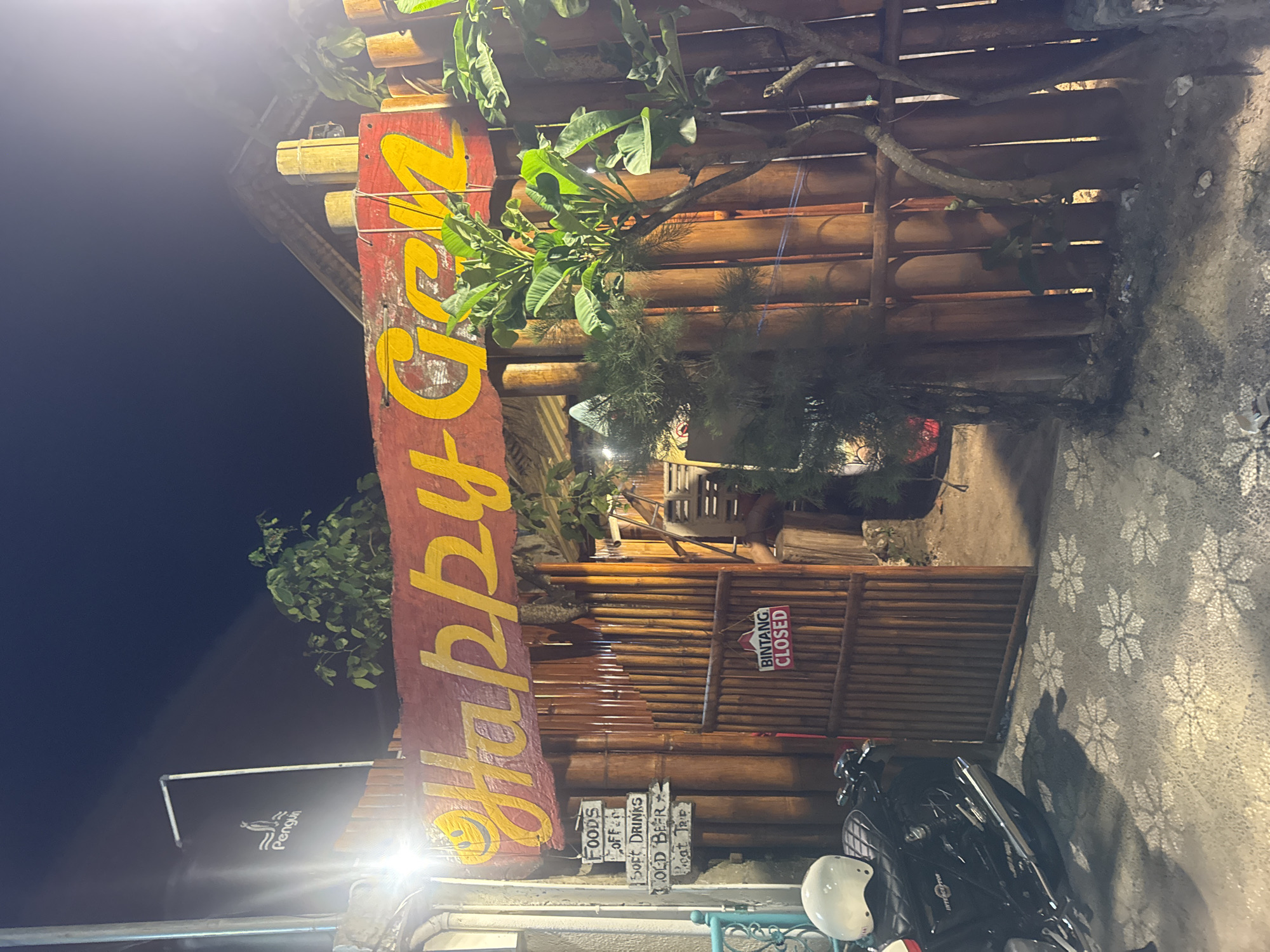 The image depicts the entrance of a restaurant in Kuta, Indonesia, at night. The restaurant's sign reads "Happy GED". The structure is primarily constructed from bamboo. A sign indicating "Bintang CLOSED" hangs near the entrance. Below, a hand-painted sign lists offerings, including "Foods," "Soft Drinks," and "Cold Beer." A motorcycle with a white helmet is parked outside. The pavement is decorated with a floral pattern. Green foliage is visible atop the structure.