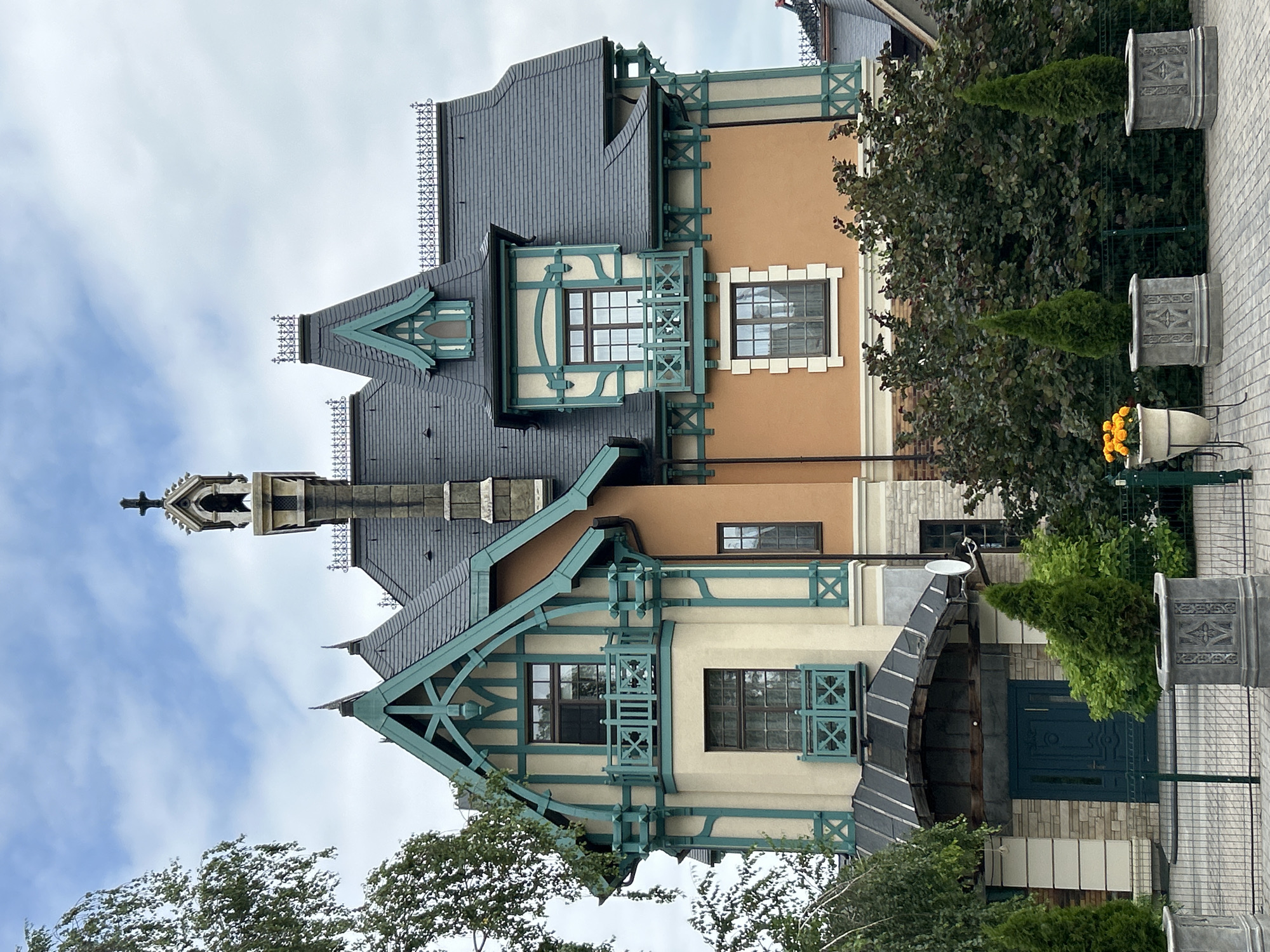 The image shows a large, multi-story house with a distinctive architectural style, possibly European-inspired, located in Khryashchevka, Russia. The house has a dark, steeply pitched roof with decorative finials. The facade is a combination of light beige and orange sections, accented with teal trim around windows, balconies, and structural elements. Several windows are visible, some with balconies featuring teal railings. The entrance of the house is covered by a dark, angled awning leading to a teal double door set within a stone-clad entryway. In front of the house, there is a paved walkway bordered by planters. Two conical evergreen trees are in square, ornate planters, and another planter to the left holds bright orange flowers. Behind the trees, a dense hedge of dark green foliage lines the property. The sky is partly cloudy. To the left of the house, a portion of a large tree with green leaves is visible. No people are present in the image.