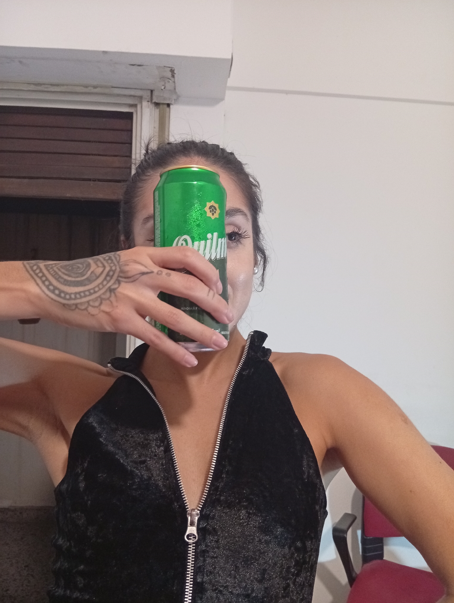 A woman is holding a green and silver Quilmes beer can in front of her face. She has a tattoo on her left hand and is wearing a black velvet top with a silver zipper. The scene is indoors with a white wall and a closed wooden door in the background. There is a red chair to the right. Buenos Aires, Argentina is not visible in the image.