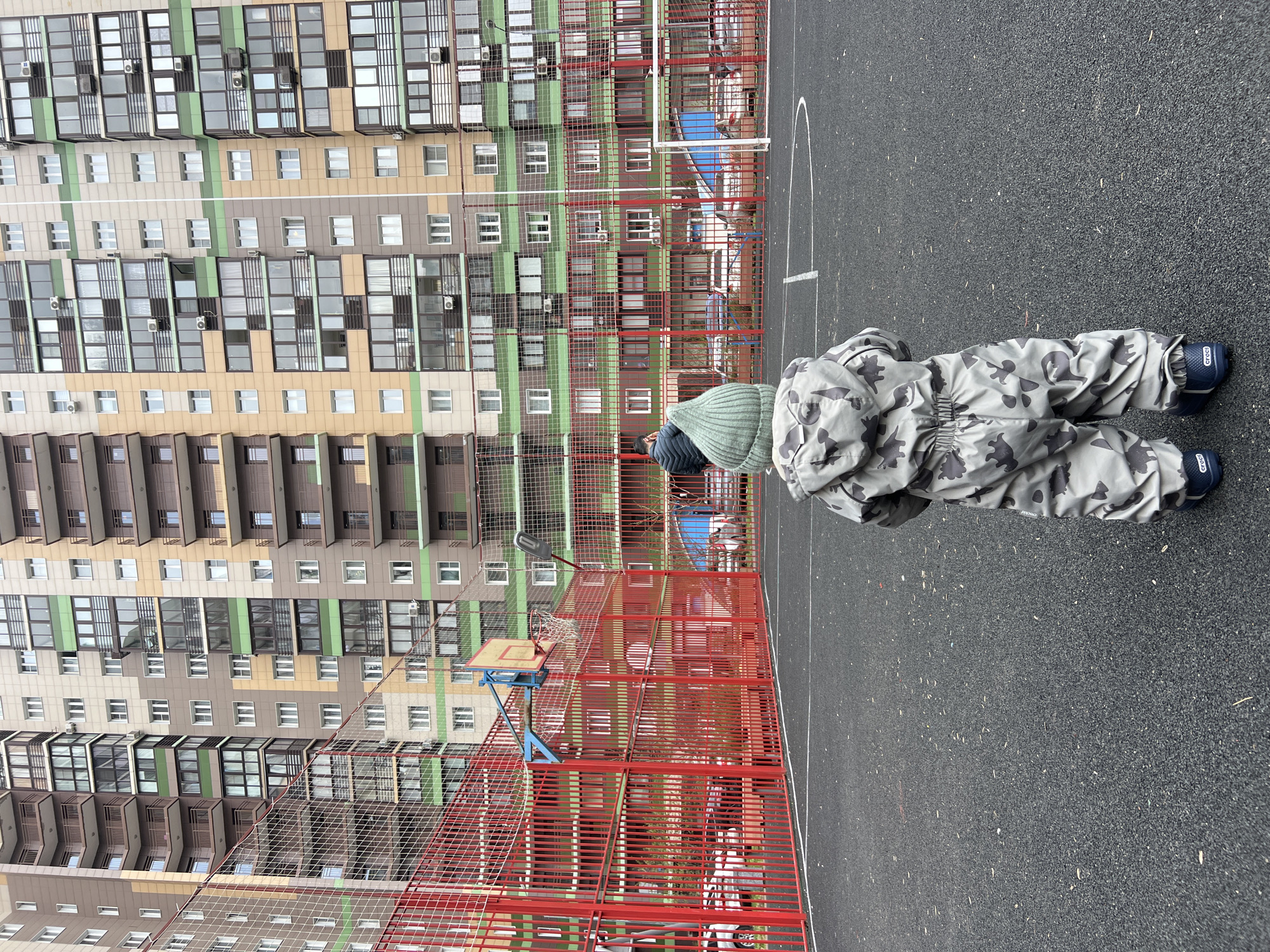 The image, rotated 90 degrees counter-clockwise, captures an outdoor scene featuring a person on a sports court with a large residential building in the background. The central subject is a person, likely a child, dressed in a grey camouflage-patterned full-body suit and a green knitted hat. Seen from behind, they are bent over, looking towards the ground. Their feet are covered in blue shoes with the "Crocs" brand name clearly visible on the heels.
The setting is a dark grey, textured asphalt sports court with white markings partially visible, suggesting a basketball court. A red metal fence, topped with a lighter mesh netting, runs across the mid-ground, separating the court from the background. A basketball hoop with a blue support structure, a yellow backboard, and a white net is prominent on the left side of the fence. A street light is also attached to the fence.
Dominating the background is a modern, multi-story residential building with a facade featuring horizontal stripes of beige, light green, and brown sections. Numerous windows, some with reflective surfaces, and balconies are visible across its many floors. Several air conditioning units are mounted on the building's exterior. The sky appears overcast, indicating a cloudy day, and there are no strong shadows. The overall scene suggests a residential area in an urban environment, specifically in Khimki, Russia. The person's exact activity is not clearly discernible beyond bending down.