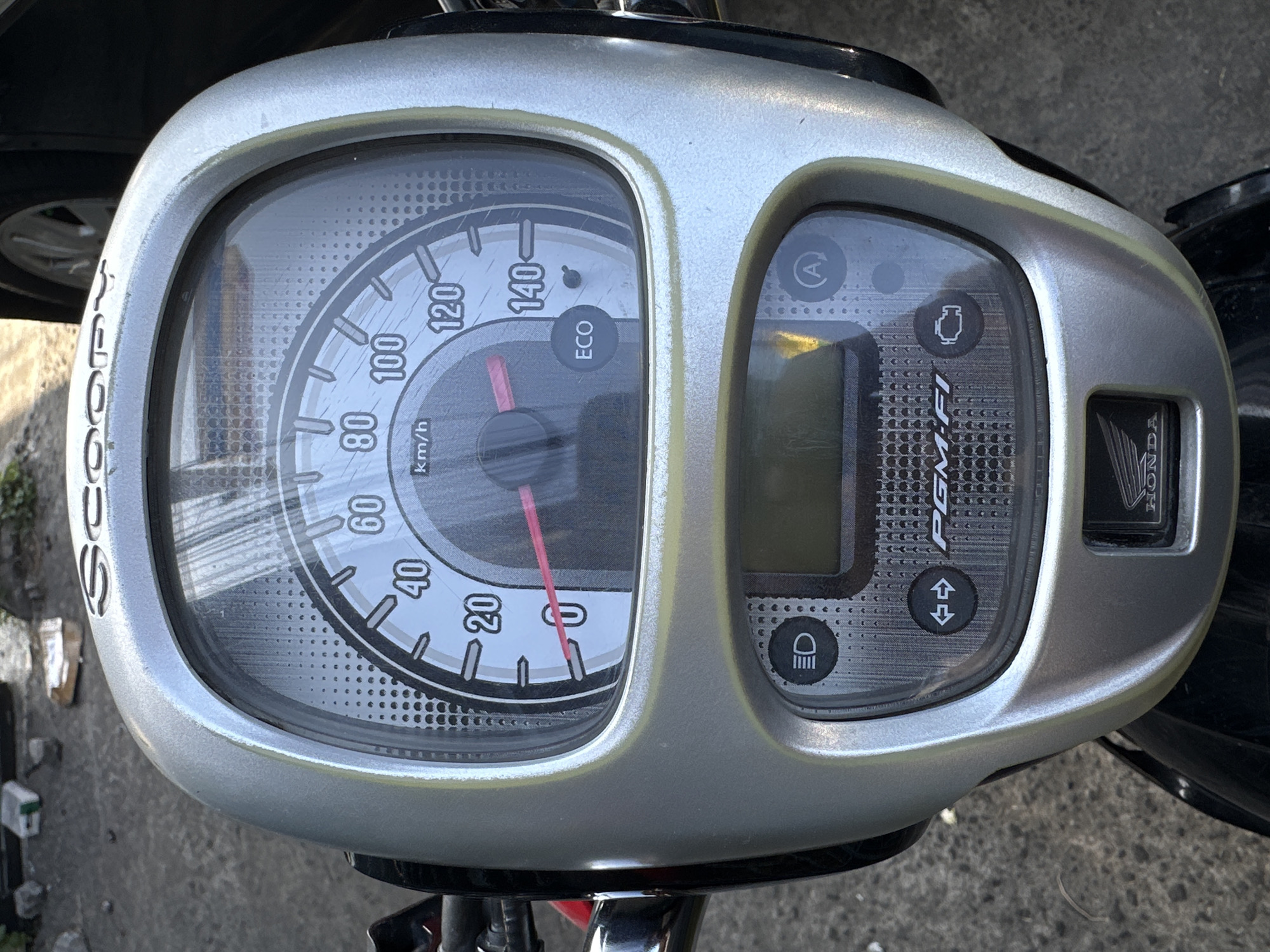 The image shows a close-up of a silver Honda Scoopy motorcycle dashboard. The dashboard features a speedometer with markings up to 140 km/h, a fuel gauge, and various indicator lights. The speedometer needle is pointing just above the 0 mark, indicating the motorcycle is stationary. Below the speedometer is a digital display and several buttons, including one with a headlight icon and another with a double-arrow icon. The "PGM-FI" logo is visible on the lower part of the dashboard, signifying fuel injection technology. The Honda logo is also present on the lower section. The background is blurred and appears to be an outdoor setting with a concrete surface and some debris, consistent with a street or parking area in Parangjoro, Indonesia. The lighting suggests it is daytime, with sunlight casting shadows. There are no people visible in the image.