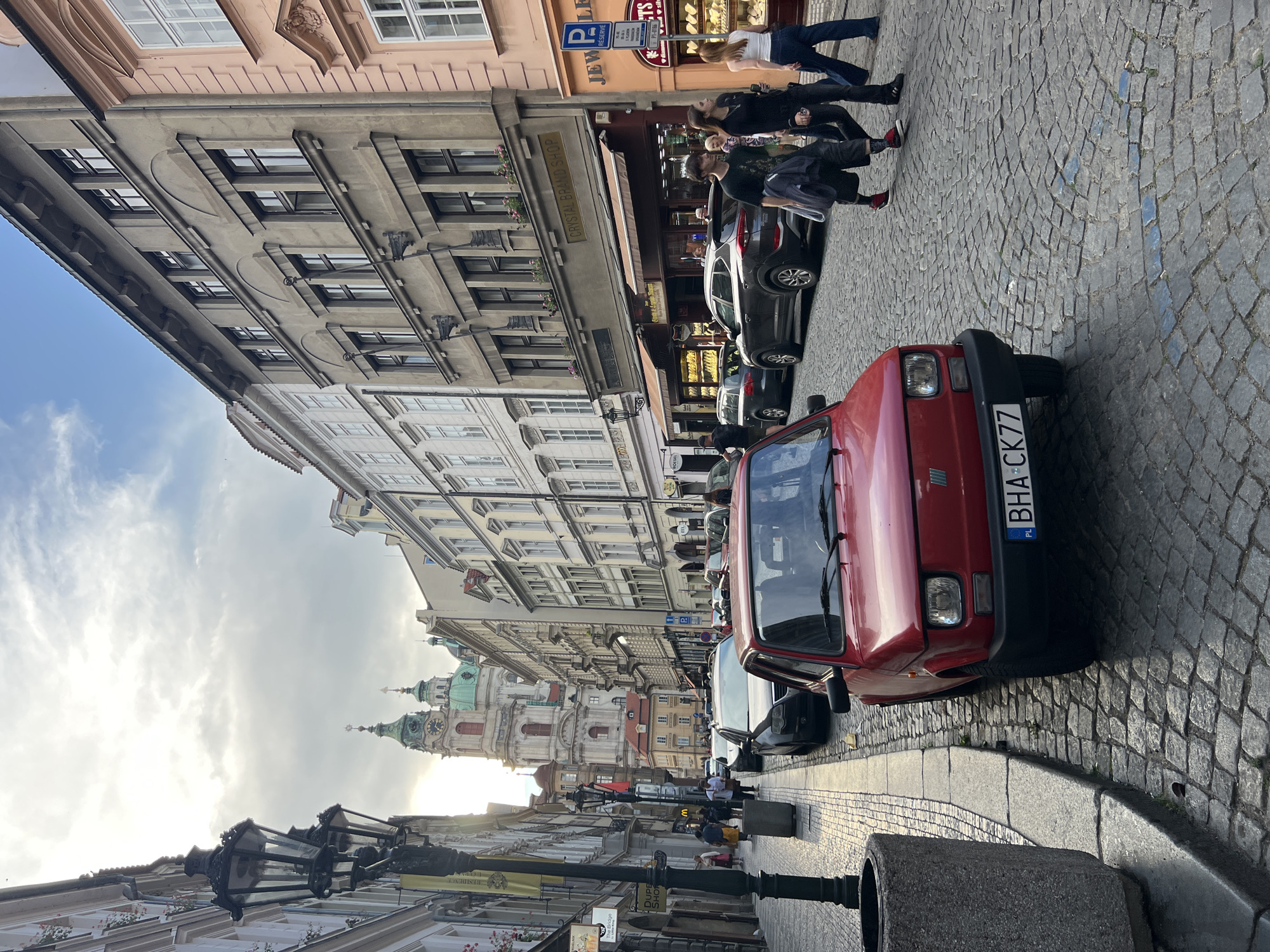 This is a medium-angle, eye-level shot of a cobblestone street in a European city. In the foreground, a small red car is parked on the left side of the street, facing away from the viewer. The car has a license plate that reads "BHA CK77". To the left of the car is a large, gray, cylindrical trash can.
The street is lined with buildings of varying architectural styles and colors, typical of an old European city. The buildings have multiple stories and are adorned with windows and decorative elements. Some of the buildings appear to house shops with awnings. In the background, a large, ornate church or cathedral with green domes and a clock tower is visible.
Several people are walking on the sidewalk on the right side of the street. They are dressed in casual clothing. Some are walking individually, while others are in small groups. The sky is partly cloudy, with sunlight breaking through. The overall impression is a busy urban scene in daylight.