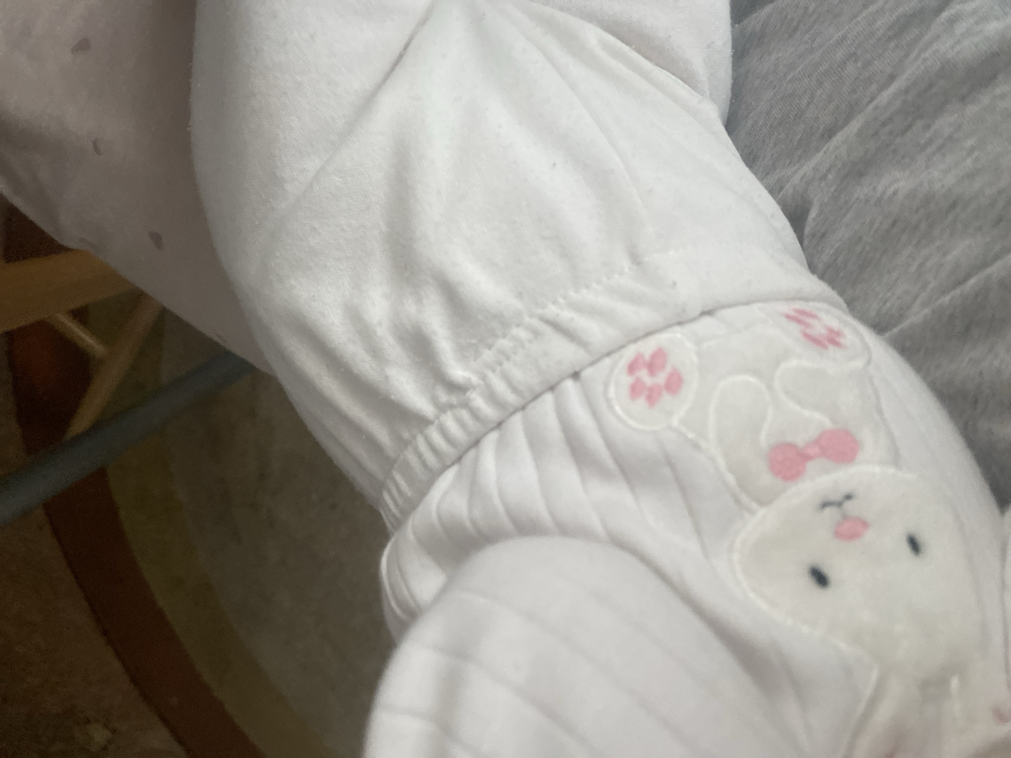 The image shows the arm and lower body of an infant dressed in white pajamas. The pajamas feature a white bunny design with a pink bow and pink paw prints. Part of a wooden chair can be seen in the background, along with what appears to be a gray blanket or garment. No information is provided to connect this image to Blagoevgrad, Bulgaria.