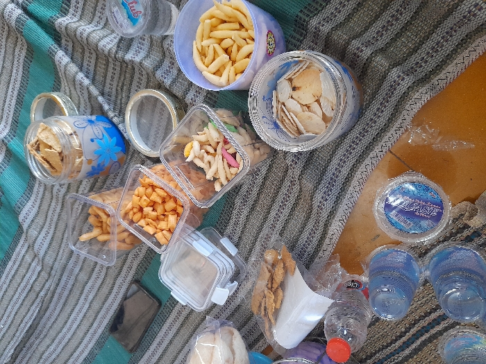 The scene depicts an assortment of snacks and beverages laid out on a striped woven mat, composed of alternating horizontal bands of grey/beige and teal/turquoise. A light brown floor surface is visible beneath the mat.
The items present include:
* Multiple clear plastic square containers holding various snacks: one with golden-brown elongated items, one with small orange square-shaped items, and another with mixed light-colored, irregularly shaped snacks with colorful specks.
* A clear glass mug containing light-brown, irregular-shaped flat crackers.
* A light-purple plastic bowl filled with numerous light-yellow, elongated, curved snacks.
* A clear plastic jar holding multiple round, thin, light-colored chips or crackers.
* A plastic-wrapped package of flat, irregular-shaped crackers resting on a white sheet of paper or cardboard.
* A partially visible white, round item contained within a clear plastic bag.
* Several clear plastic water bottles with red caps, one partially consumed.
* Multiple stacks of clear plastic cups.
* A single clear plastic cup containing water, labeled "Club."
* A sealed clear plastic package containing multiple small, pre-filled water cups with blue and white branding.
* A dark-colored smartphone, screen-down, positioned on the mat.
* A metallic ring, possibly a container lid, located near the center of the arrangement.
The arrangement suggests a casual gathering or communal snack setting in Kebomas, Indonesia.