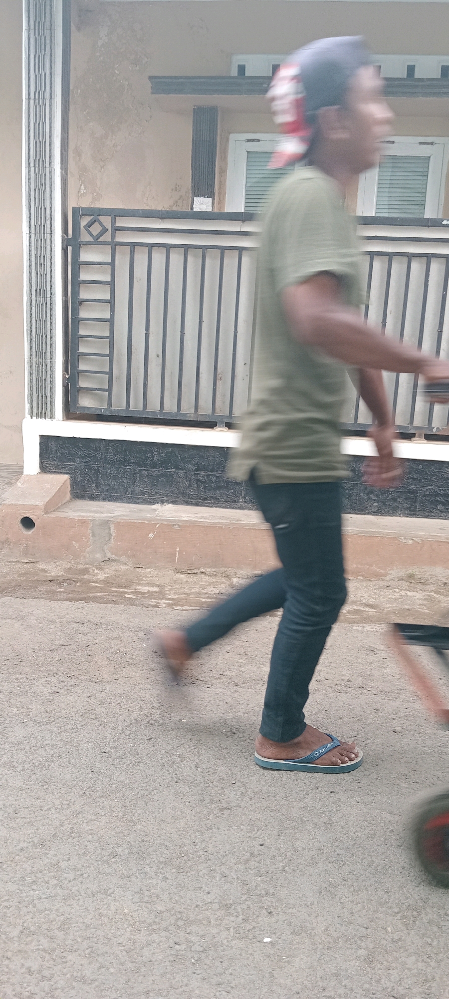The image captures a motion-blurred view of a person walking on a paved street in Tasikmalaya, Indonesia. The individual, wearing a green t-shirt, dark pants, and flip-flops, is moving from left to right, with their right leg extended forward. Behind them, a portion of a building is visible, featuring a dark-colored textured base and a lighter upper section. A black metal fence with a diamond pattern separates the building from the street. The environment appears to be an outdoor urban setting.