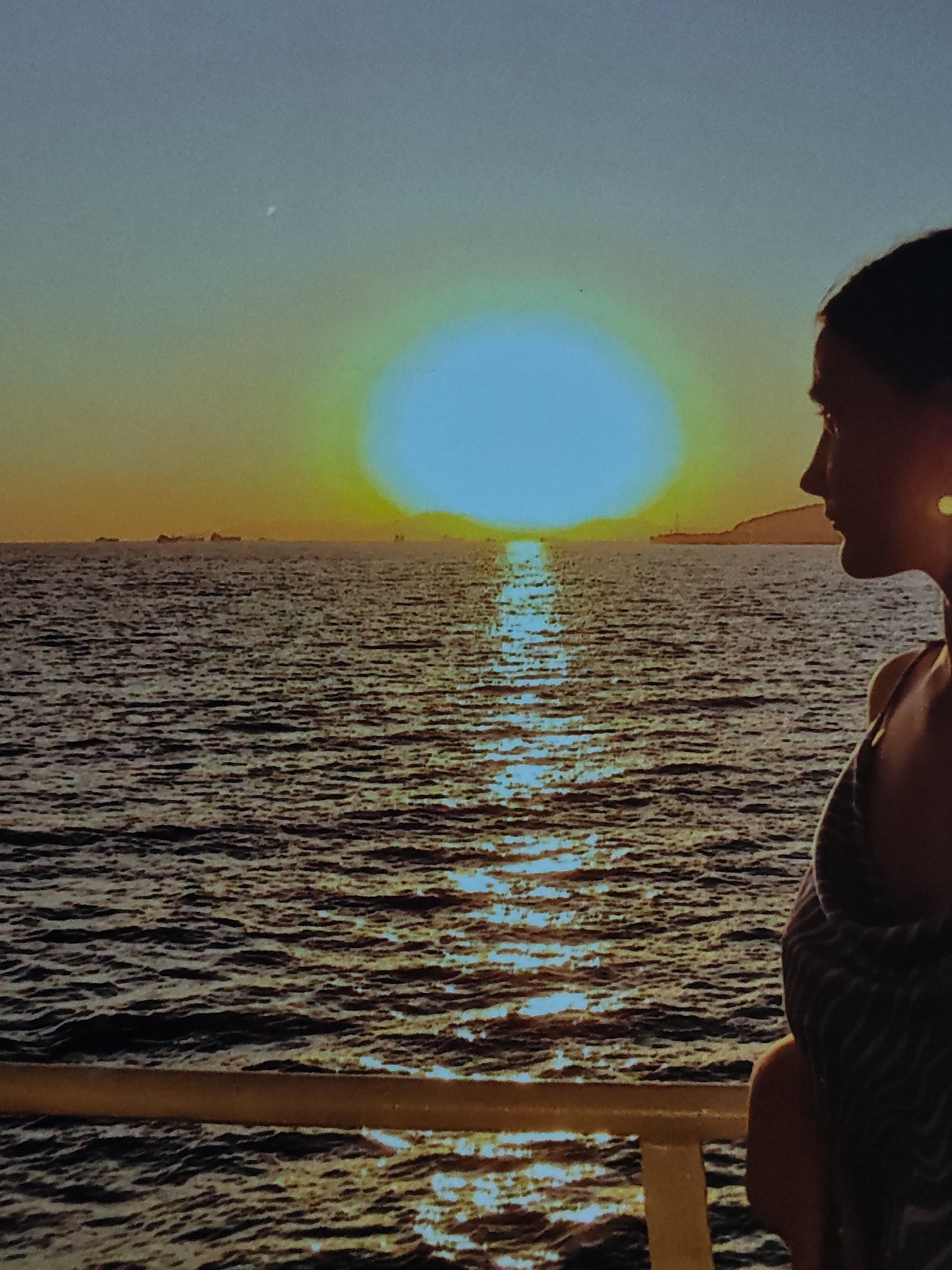 An eye-level shot captures a woman in profile, her face turned towards a brilliant sunset over a calm sea. The sun, a large, glowing orb of blue and yellow, is positioned slightly left of center, casting a shimmering reflection across the water's surface. In the distance, silhouetted ships are visible on the horizon. The woman, with dark hair and a light-colored earring, wears a striped garment. She stands at the edge of what appears to be a boat or pier, with a railing visible in the foreground. The sky transitions from a pale blue at the top to warm yellow and orange hues near the horizon.