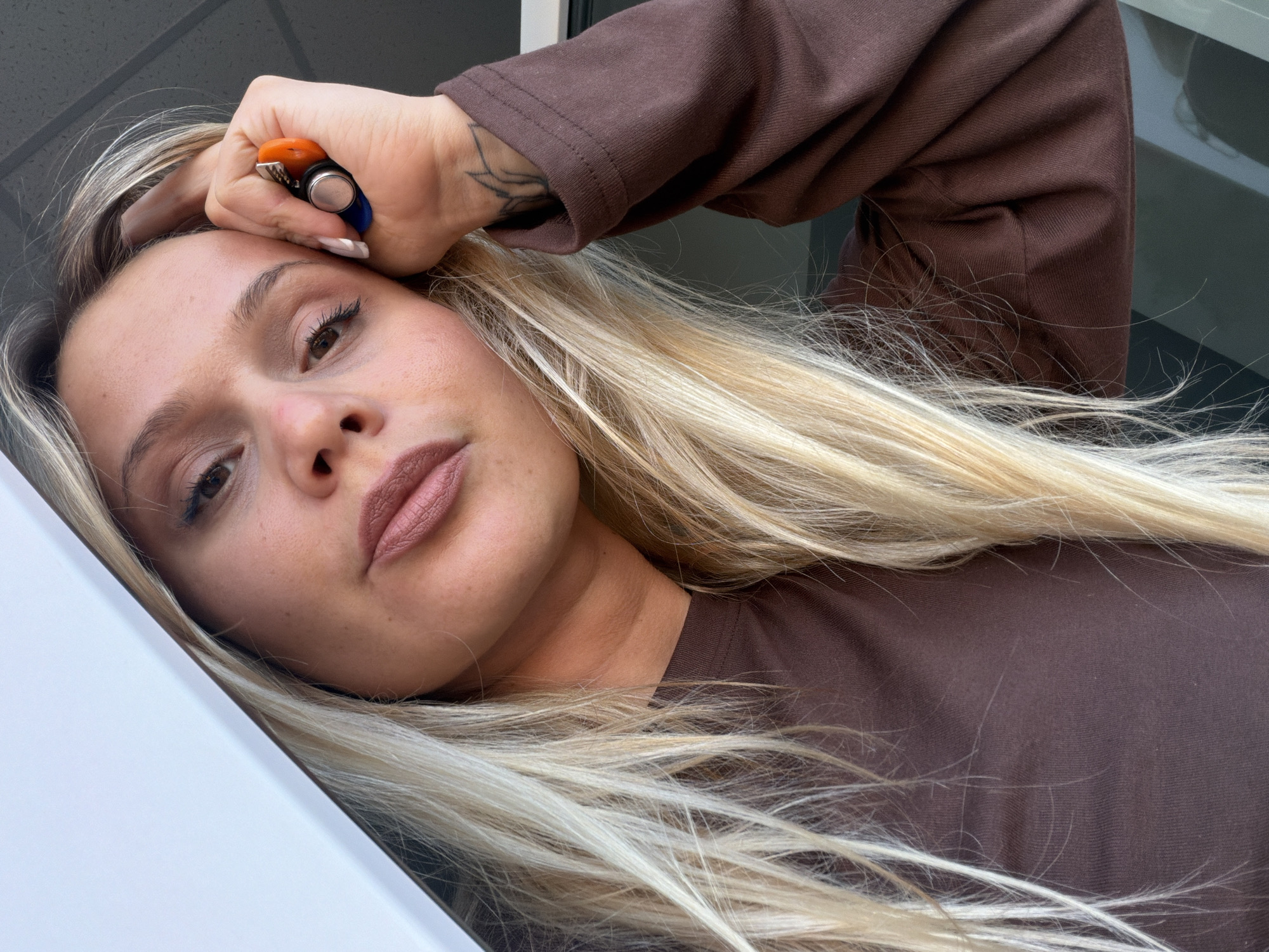 A woman with long, blonde hair is positioned close to the camera, her head tilted. She is wearing a brown long-sleeved shirt. Her hand, adorned with a tattoo, rests on her forehead. The scene includes a glimpse of a white surface and a grey reflective surface. The woman holds what appears to be a multi-colored object, possibly a lighter or similar device, between her fingers. There is no explicit information in the image to confirm the location in Sarapul, Russia.