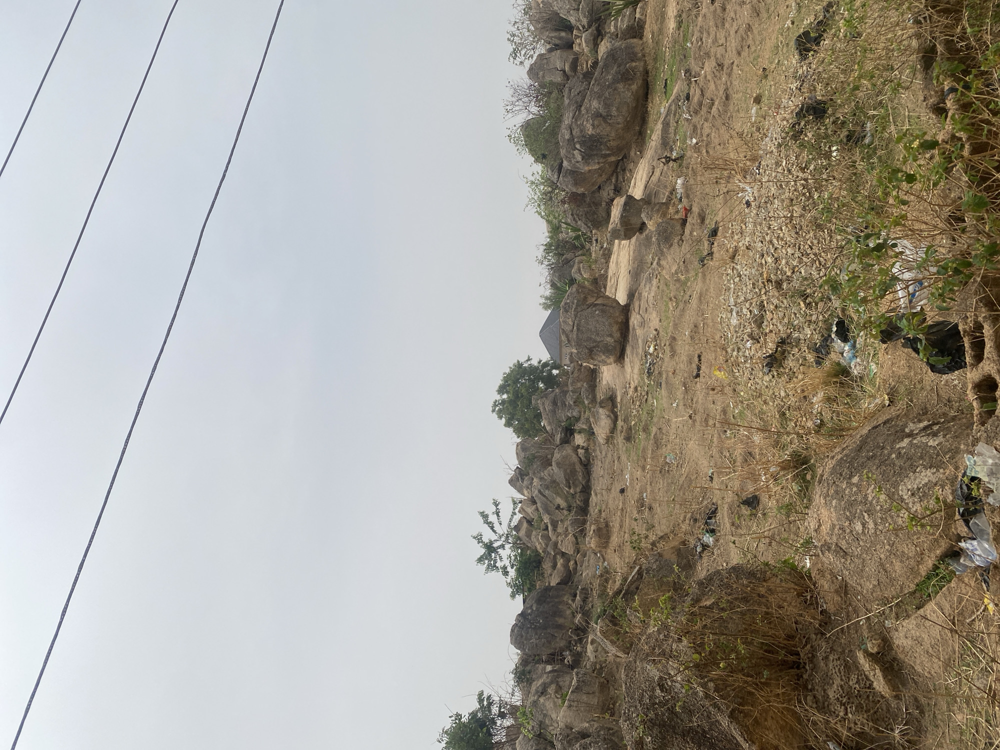 The image shows a rocky, uneven terrain with sparse vegetation in what appears to be an outdoor area in Barakin Akawo, Nigeria. A group of large boulders and smaller rocks are scattered across the landscape. A small structure with a dark roof is partially visible among the rocks and trees. Litter and debris are present near the rocks. Power lines run across the upper left portion of the frame. The sky is overcast.