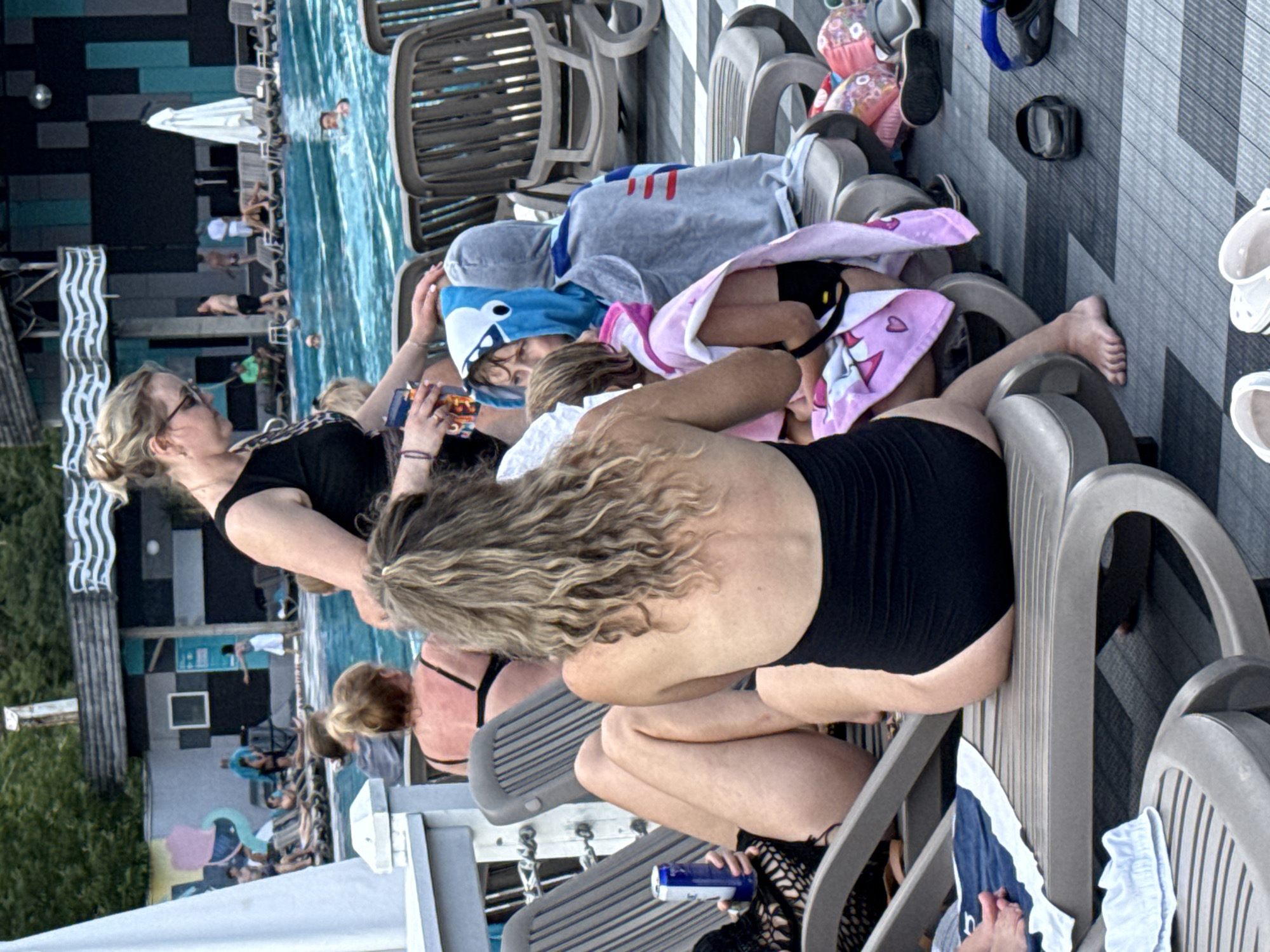This image depicts a poolside scene with several individuals. In the foreground, a woman with blonde, wavy hair is sitting on a lounge chair, wearing a black swimsuit. To her left, another woman is partially visible, also on a lounge chair and wearing a black swimsuit. Two children are seated on lounge chairs in the middle ground. One child is wearing a blue hooded towel with a shark design, and the other is wearing a pink hooded towel. A woman, presumably an adult, is standing behind them, holding a drink. In the background, a swimming pool is visible with several people in the water. Lounge chairs are arranged around the pool, and a building with a dark facade and windows is situated behind the pool. The overall environment suggests a resort or public pool area. The location is identified as Массандровский поселковый совет, Ukraine.