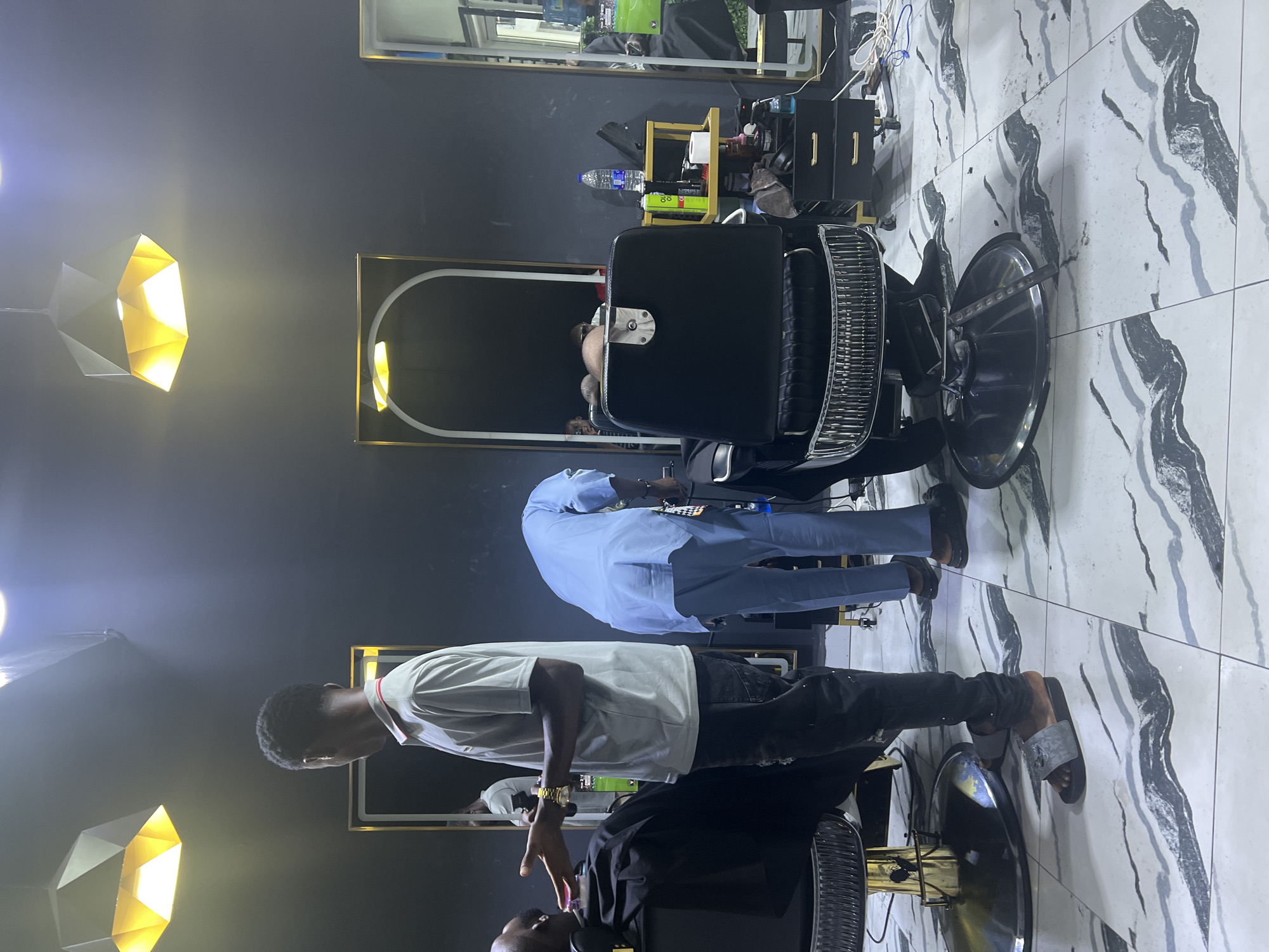 A barber shop in Lugbe, Nigeria features a customer in a black barber chair receiving a haircut from a barber wearing a white t-shirt and black jeans. Another individual in a light blue shirt stands nearby. The shop has black walls with framed mirrors, marble-patterned tile flooring, and modern lighting fixtures. A shelf unit with various barbering supplies is also visible.