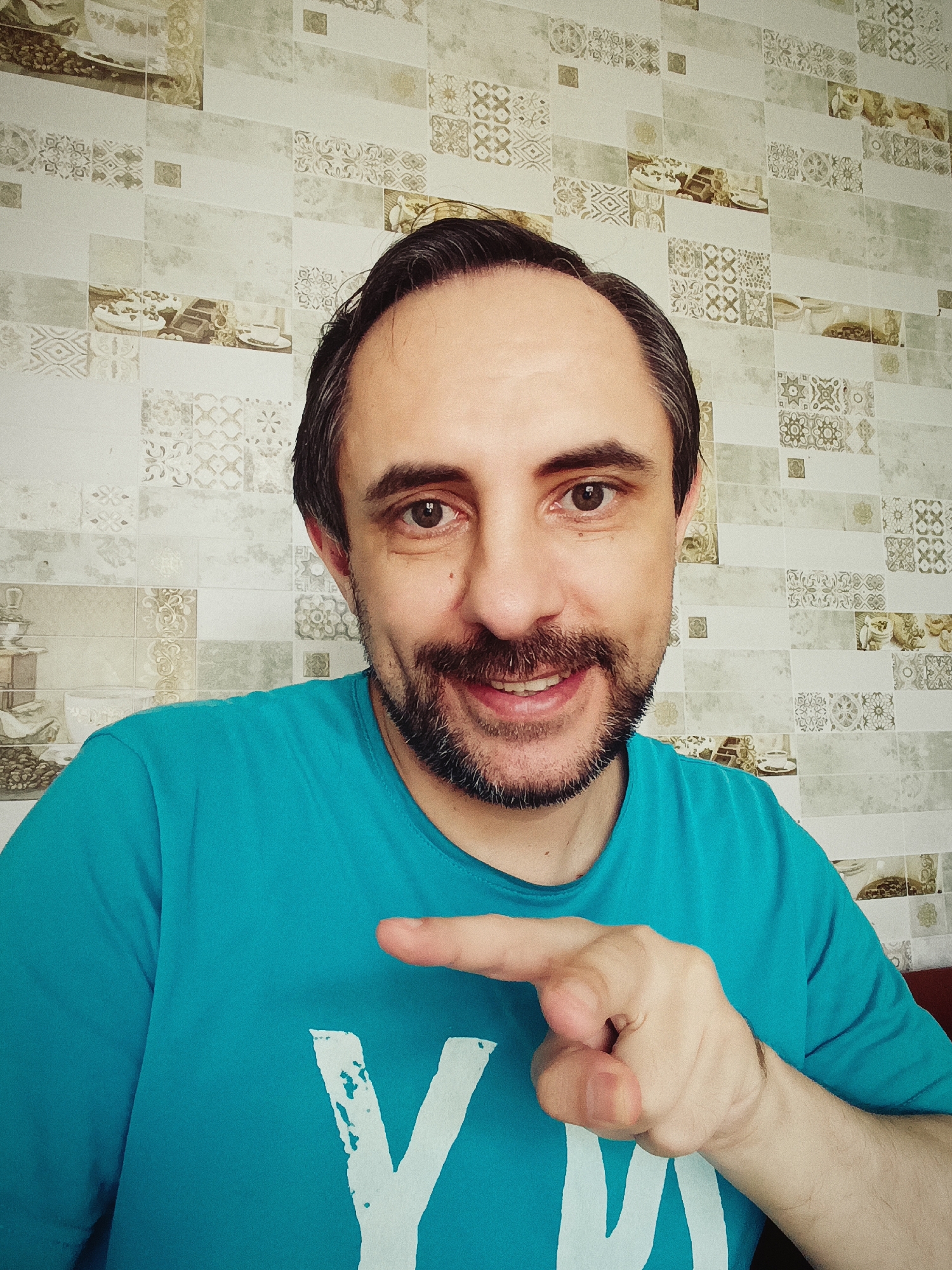 A head and shoulders, waist-up shot shows a man with a beard and short, dark hair, smiling into the camera. He is wearing a turquoise t-shirt with a white, abstract design. His right arm is bent, and he is pointing his index finger toward the viewer. The man is positioned in front of a wall covered in decorative tiles. The background shows a close-up of the man's face and chest, as well as the patterned wall. The image appears to be a selfie. There is no specific indication of the location being the city of Izmail, Ukraine, in the visual information provided.