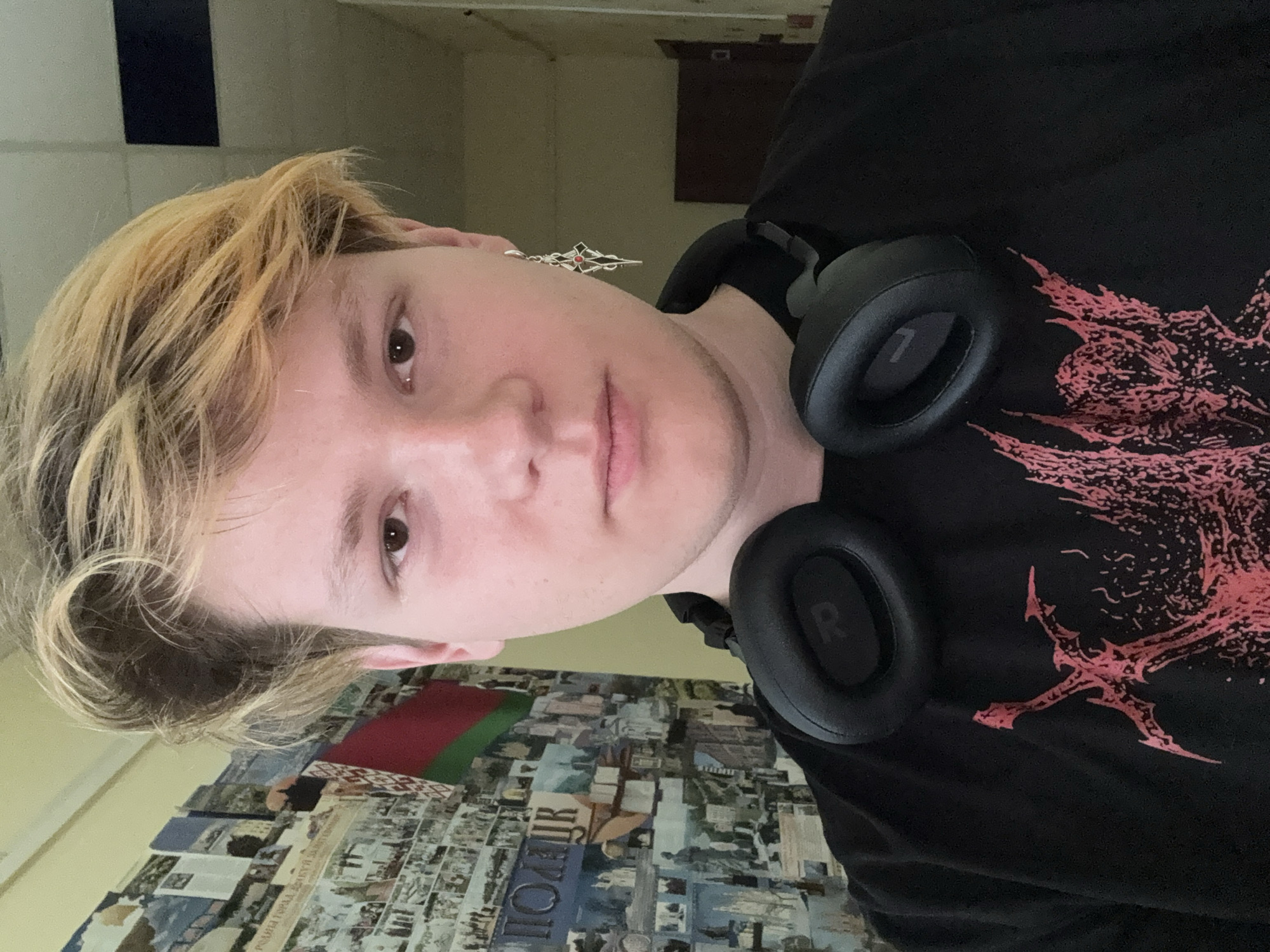 The image is a selfie taken indoors, likely in a hallway, featuring a young person with blonde-highlighted hair and a black t-shirt. They are wearing black over-ear headphones around their neck and a silver earring with a gothic design in their left ear. The background on the left wall is covered with a collage of pictures and what appears to be a Belarusian flag. The lighting is dim, casting shadows on the wall behind the subject. The overall setting suggests a casual, personal space within the Ветрынскі сельскі Савет, Belarus.