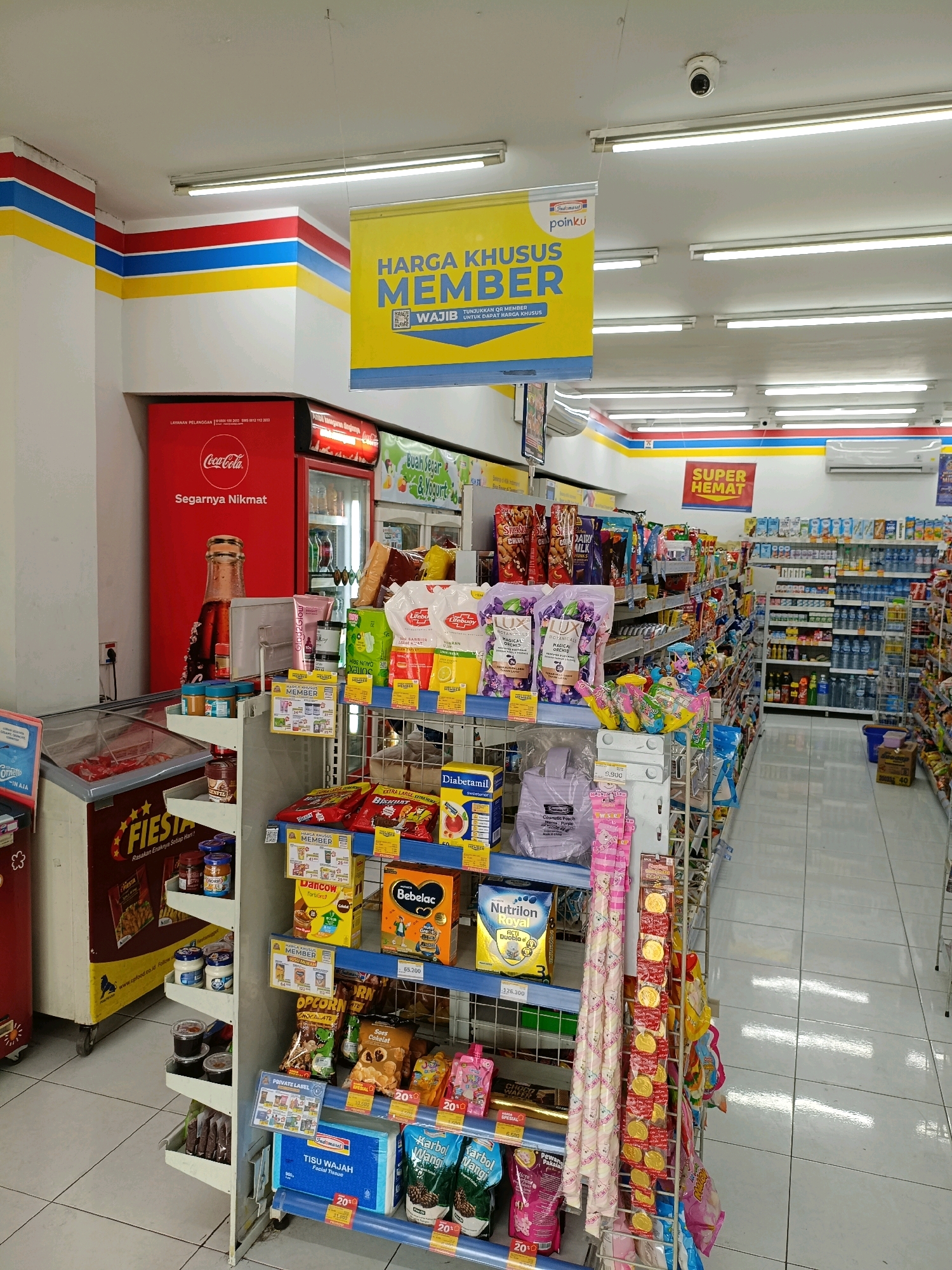 The image shows the interior of an Indomaret convenience store in Maguwoharjo, Indonesia. The store is brightly lit with fluorescent lights and features aisles stocked with various consumer goods. A large yellow banner hangs from the ceiling, advertising "HARGA KHUSUS MEMBER" (Special Member Price) with the logo "pointku". In the foreground, shelves display products such as snacks, drinks, personal care items, and household goods. A Coca-Cola refrigerator is visible on the left, and an air conditioning unit is mounted high on the wall. The floor is tiled. There are no people visible in the image.
