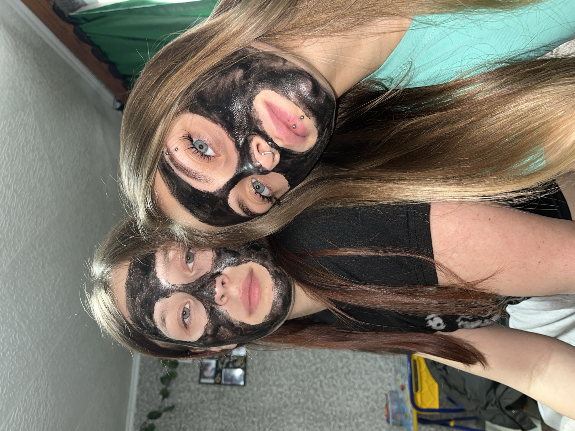 Two individuals, presumably females, are depicted from a close-up, overhead perspective. Both have dark, paste-like facial masks applied, leaving only areas around their eyes and lips visible. The upper individual has blue eyes and facial piercings near the eye and lip. The lower individual has green eyes. Long hair of varying shades of brown frames both faces. The upper individual wears a teal top, and the lower individual wears a black top. The background includes a bed, a wall with framed photos, and miscellaneous household items. There is no indication of the city of Фоки, Russia.