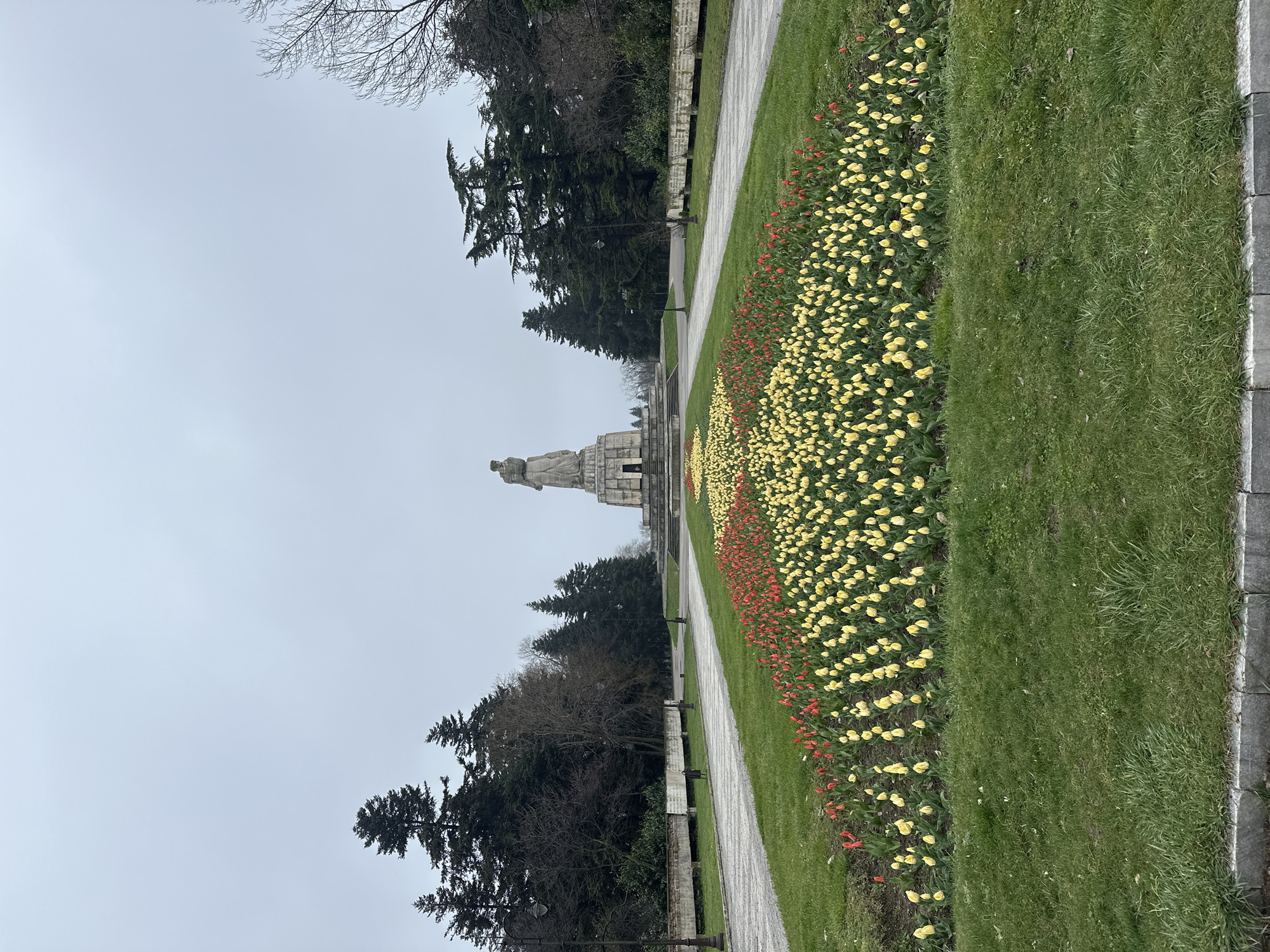 The image shows a straight walkway in a park leading toward a large monument. The walkway is flanked by flowerbeds filled with yellow and red tulips and stretches of green grass. The trees are on the left side of the walkway. The monument is light-colored stone and has a figure atop it. The sky is overcast. This is most likely the Monument of the Soviet Army in Varna, Bulgaria.