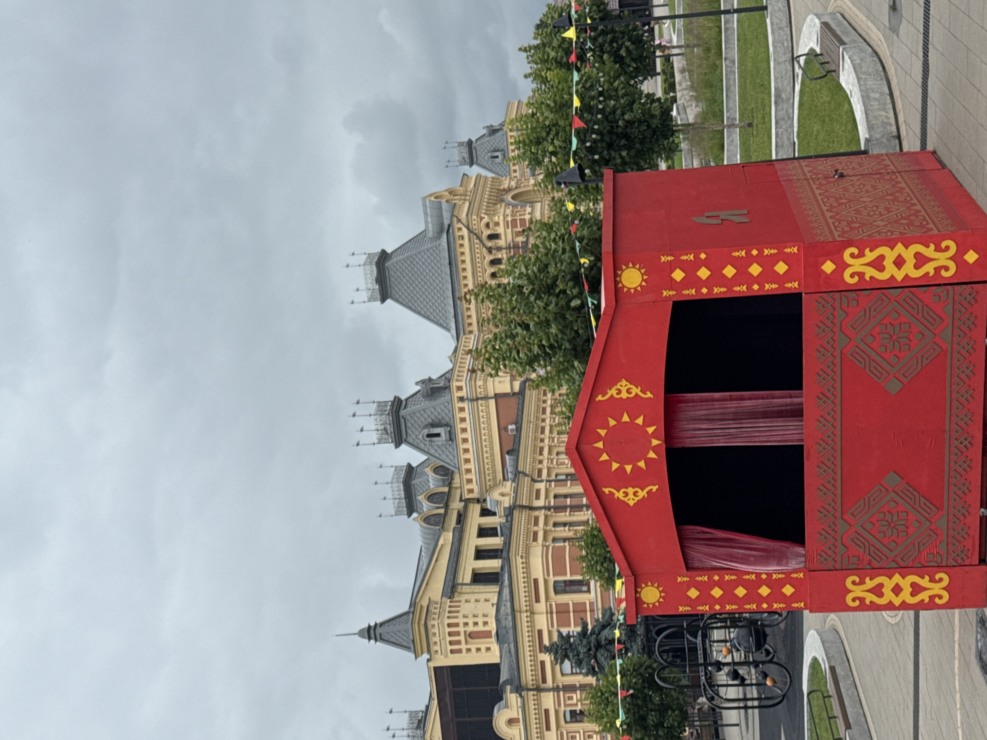 An overcast day in Nizhny Novgorod, Russia, features a small, bright red puppet theater in the foreground. The theater is adorned with yellow and brown decorative patterns, including sun-like motifs and geometric designs. Behind the theater, a large, ornate building with yellow and red brick accents and a grey tiled roof stands prominently. Trees and decorative flags add to the outdoor setting, with a paved walkway and a curved bench visible in the lower right portion of the image.