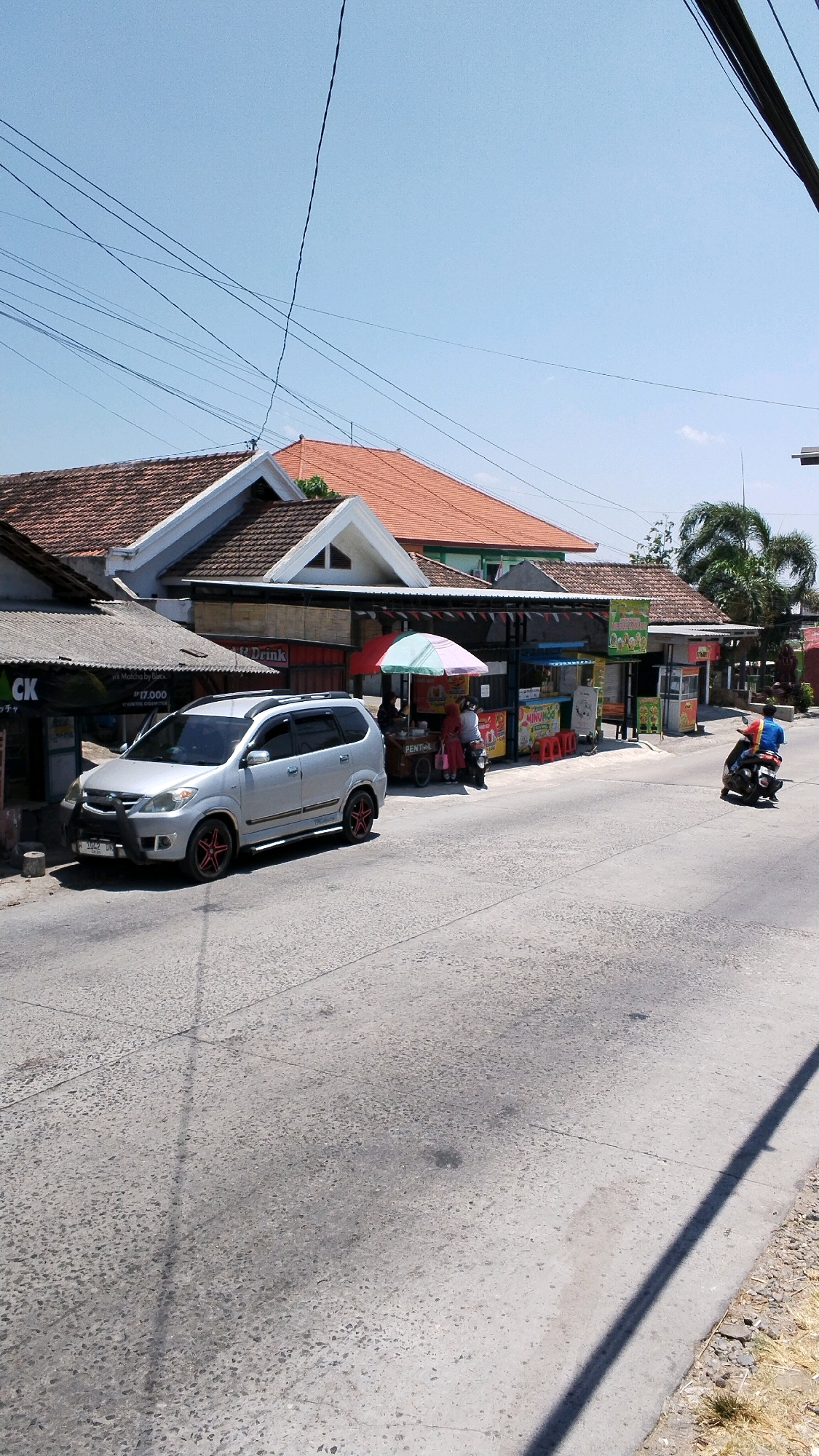 The image depicts a street scene in Pulorejo, Indonesia, on a clear, sunny day. The foreground shows a gray asphalt road with visible cracks and some debris. On the left side of the road, a silver minivan is parked. Behind the minivan, there are several small storefronts and stalls. One stall has a sign that reads "Matcha by Black" and displays a price of "P 17.000". Further down the street, a row of food stalls is set up, some with colorful umbrellas and awnings. A few people are visible near the stalls, including two individuals standing by a food cart under a multicolored umbrella. To the right of the food stalls, a person in a blue shirt is riding a black motorcycle away from the camera. Palm trees are visible in the background, suggesting a tropical or subtropical climate. Overhead, several electrical wires crisscross the sky, casting shadows on the scene. The buildings have terracotta tile roofs.