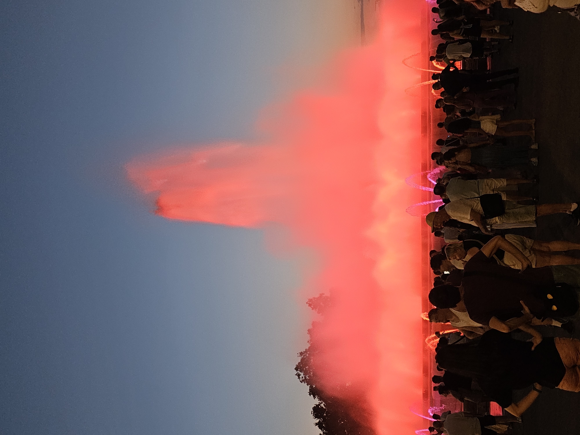 The image shows a crowd of people gathered in front of a large, illuminated fountain at dusk in Barcelona, Spain. The fountain is spraying water high into the air, and the water is illuminated with a vibrant red light, creating a dramatic effect against the darkening sky. The crowd, consisting of individuals of various ages, are standing and watching the fountain display. Some people are interacting with each other, while others are focused on the spectacle. Trees are visible in the background, silhouetted against the sky and the fountain's glow. The ground in front of the fountain appears to be a paved area. The lighting suggests it is either sunset or a controlled lighting display taking place after dark. The scene depicts a public gathering at a landmark attraction.
