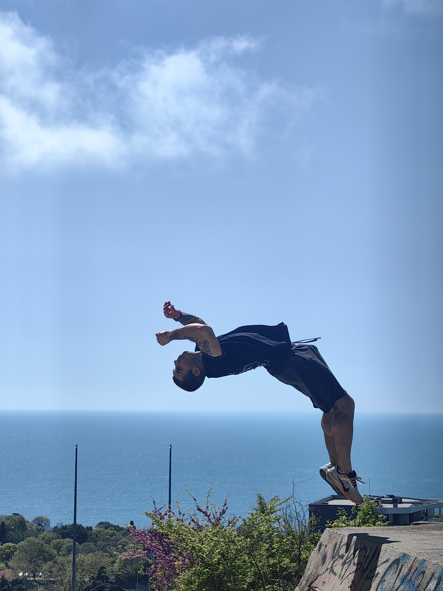 A man is performing a backflip on a concrete structure. He is wearing a dark t-shirt, dark shorts, and light-colored sneakers. His body is arched backwards, with his arms raised and legs extended downwards. The background features a bright blue sky with scattered clouds, the Black Sea, trees, and vegetation. The location is on the Asparuhovo bridge in Varna, Bulgaria.