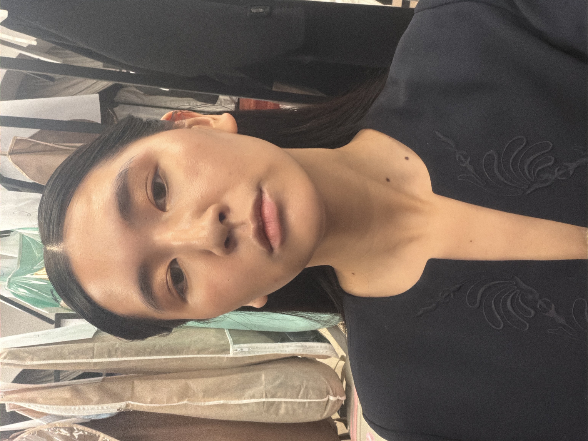 A close-up, portrait-style photograph features a young East Asian woman, depicted from the waist up. She has dark, straight hair, neatly parted in the middle and pulled back, and a clear complexion with natural-looking makeup and subtle lip color. Her expression is calm and neutral. She is wearing a dark, likely navy or black, top that features an intricate embroidered or embossed design with swirling, organic motifs on the shoulder and upper chest area. A small mole is visible on her upper right chest/shoulder.
The background shows a dense arrangement of clothing items hanging on racks. Some garments are protected by clear plastic garment bags, while others are covered in light-colored, possibly beige or brown, fabric covers, suggesting a well-organized but packed space. This setting implies a fashion-related environment, such as a showroom, a dressing room, or a backstage area, consistent with a location like Paris, France. The indoor lighting is soft and even. There is no visible text in the image. The provided image file is rotated 90 degrees clockwise.
