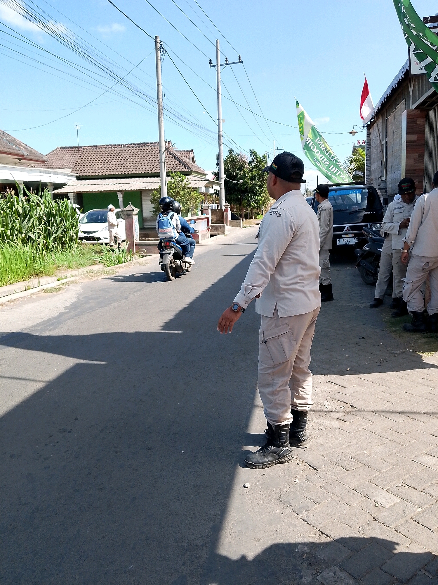 The image depicts a street scene in Banjarejo, Indonesia. In the foreground, a man wearing a light-colored uniform, a black baseball cap, and black boots is standing on a paved sidewalk, facing away from the camera. He has a watch on his left wrist. Behind him, several other individuals in similar uniforms are standing near a black SUV. A green and white flag is visible, as well as a red and white Indonesian flag. Further down the street, a white car is parked, and two people are on a motorcycle. Houses with tiled roofs line the street, and power lines are strung across the sky. The environment appears to be a sunny day with clear blue skies.