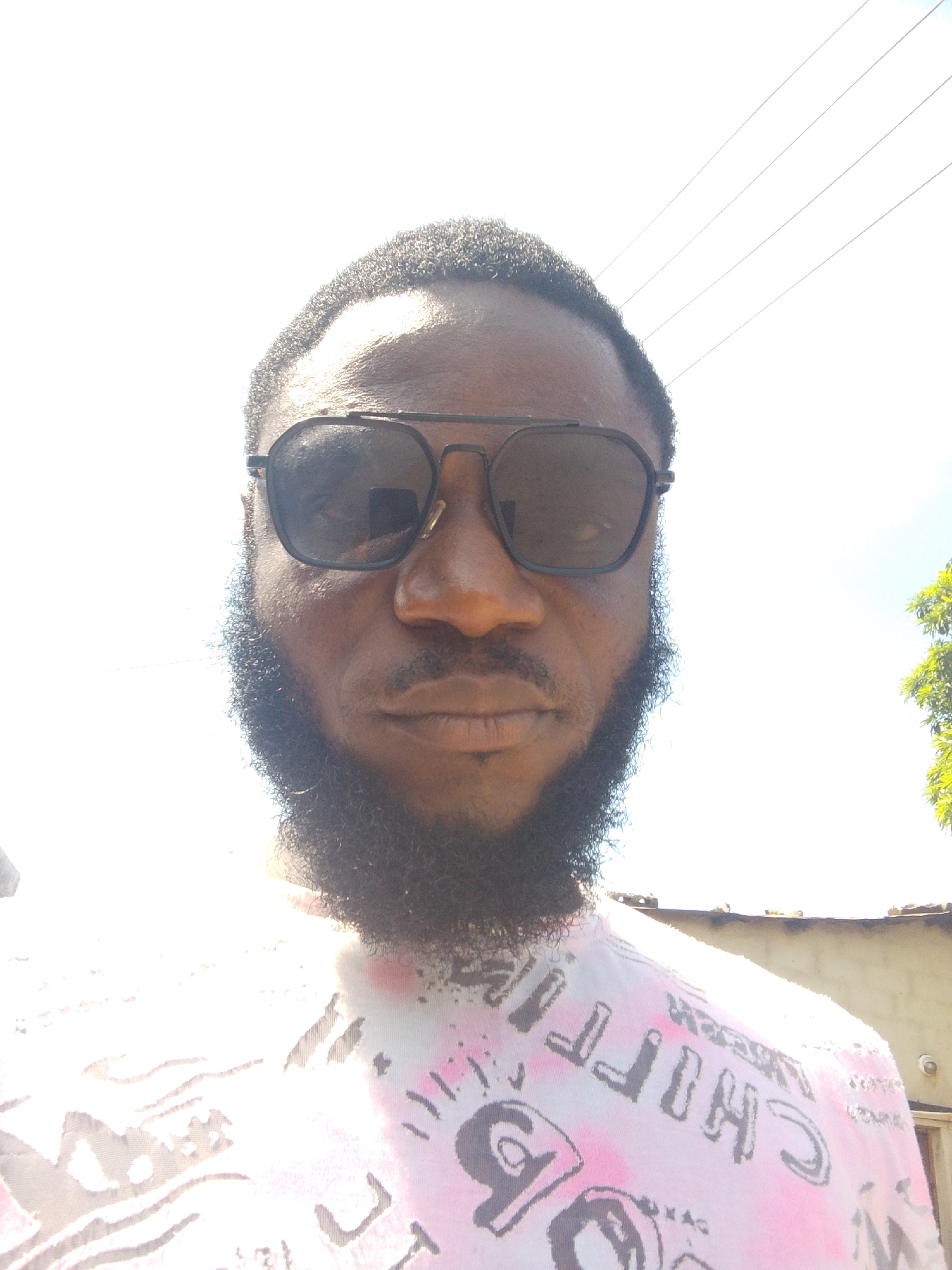 A close-up photograph shows a man with dark skin, a full beard, and short curly hair. He is wearing sunglasses with a dark frame and dark lenses. He is wearing a white patterned shirt with black and pink designs. In the background, a bright sky is visible along with the tops of buildings and green foliage. There are also overhead wires. The location appears to be outdoors in daylight. Without additional information, the specific location within Lokoja, Nigeria, cannot be determined.
