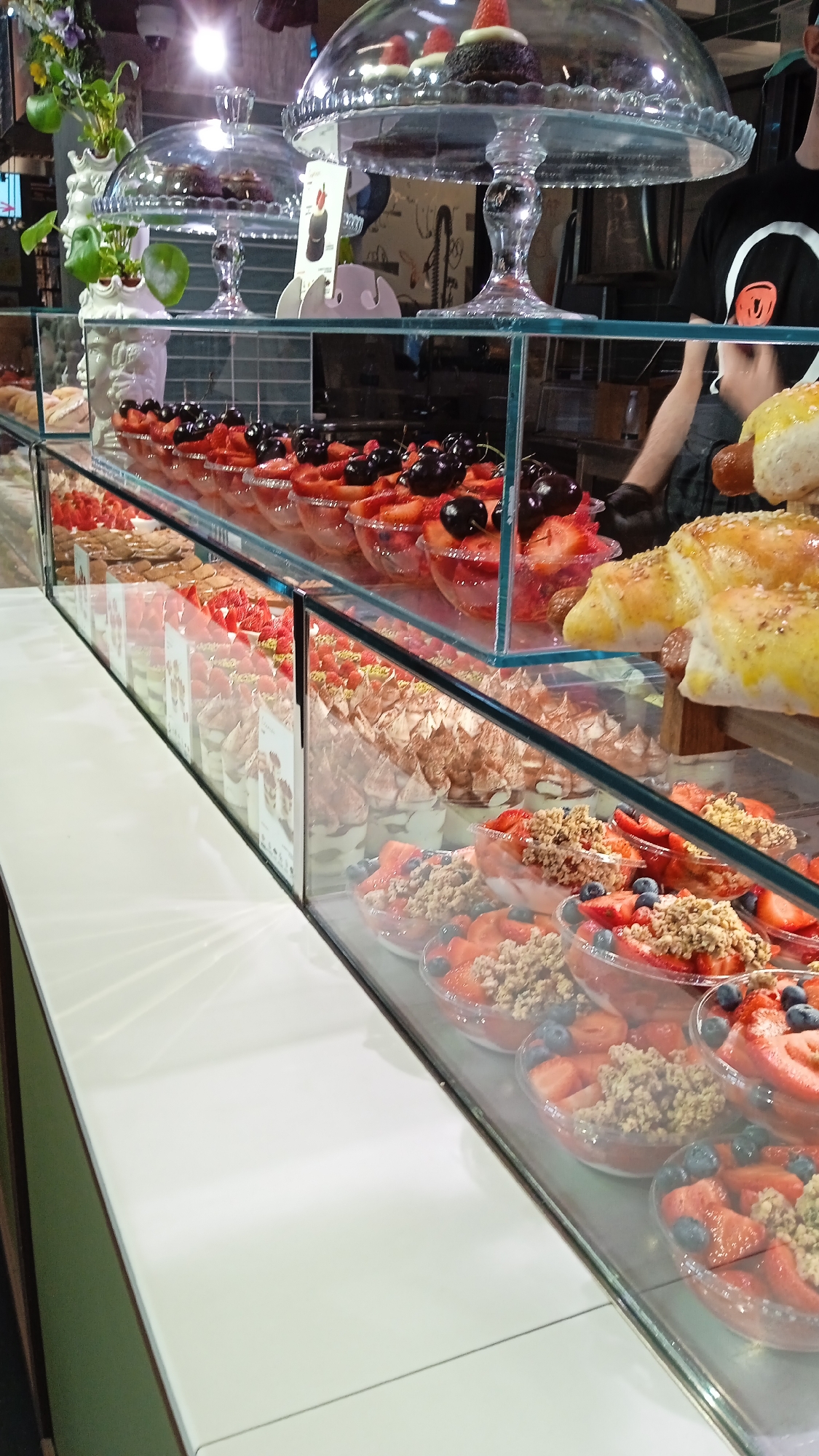 The image displays a refrigerated display case filled with various desserts, likely in a bakery or patisserie in Milan, Italy. The case is made of glass and stainless steel.
In the foreground, on the right side, there are several clear plastic bowls containing fruit salads. These salads appear to be composed of chopped strawberries, blueberries, and a topping of granola or crumbled nuts.
To the left of the fruit salads, the display case shows rows of individual desserts in clear plastic cups. These appear to be layered with cream and possibly fruit or meringue, topped with decorative elements. Some of these cups have small printed labels.
Further back and to the left, a tiered glass display stand holds small chocolate cakes, each topped with a single strawberry. Another similar stand, partially obscured, is positioned behind it.
In the background, a person wearing a black t-shirt with a circular graphic logo is partially visible. They are wearing black gloves and appear to be working behind the counter, possibly arranging or serving the desserts.
The overall environment is brightly lit, with the lighting reflecting off the glass surfaces of the display case and the polished white countertop in the foreground. The backdrop includes shelving units and some decorative elements, including a plant in a white vase on the far left. The items for sale are meticulously arranged, suggesting a professional establishment.