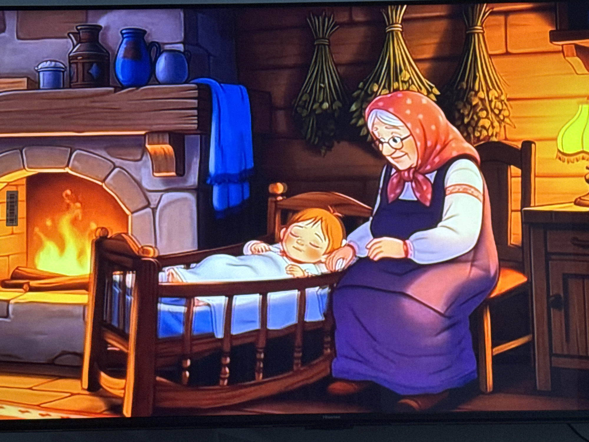 In a dimly lit, rustic interior, a grandmother figure with a red headscarf and glasses sits on a wooden chair, observing a baby sleeping in a wooden crib. The grandmother is wearing a dark blue dress with a white, embroidered top and a red tie at the neck. The baby is swaddled in a blue blanket. To the left, a stone fireplace with a roaring fire and logs is visible. Above the fireplace, a wooden mantle displays several ceramic pitchers and a small blue container. A blue cloth hangs over the mantle. In the background, two bundles of dried herbs are suspended from the wooden-paneled wall. A green lamp with a decorative base sits on a wooden cabinet to the right. The floor is tiled with a warm-toned pattern. The scene is characteristic of a cozy, traditional dwelling. The prompt's mention of "Chardara, Uzbekistan" is an irrelevant detail not supported by the visual content of the image.