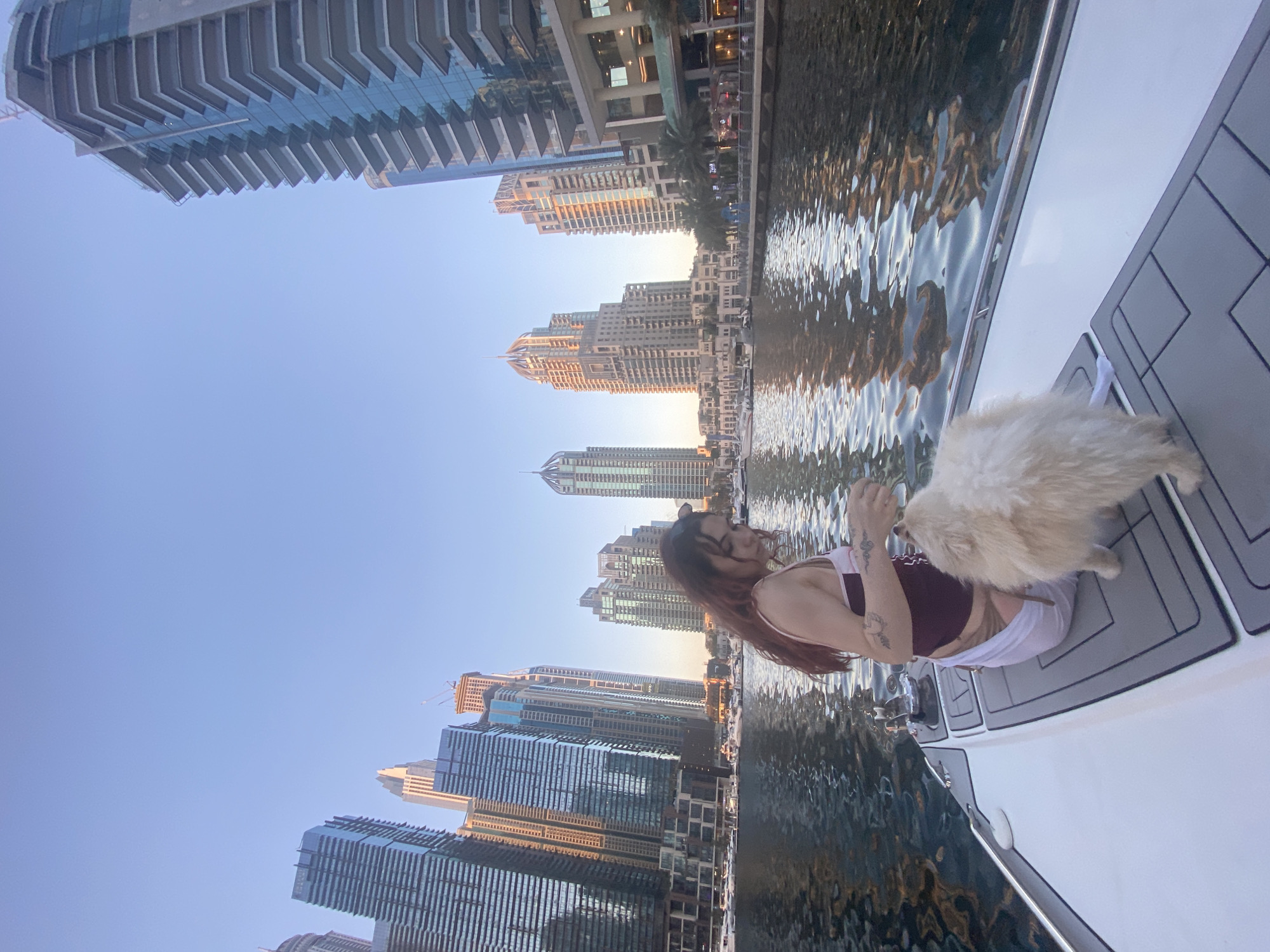 A person is on a boat in the Dubai Marina canal in Dubai, United Arab Emirates. The person has long hair, and is wearing a burgundy tank top, and white bottoms. A white fluffy dog is standing near them on the boat deck. High-rise buildings are visible on either side of the canal. The buildings are a mix of modern glass and concrete structures. The water in the canal reflects the buildings and the sky. The sky is a clear blue.