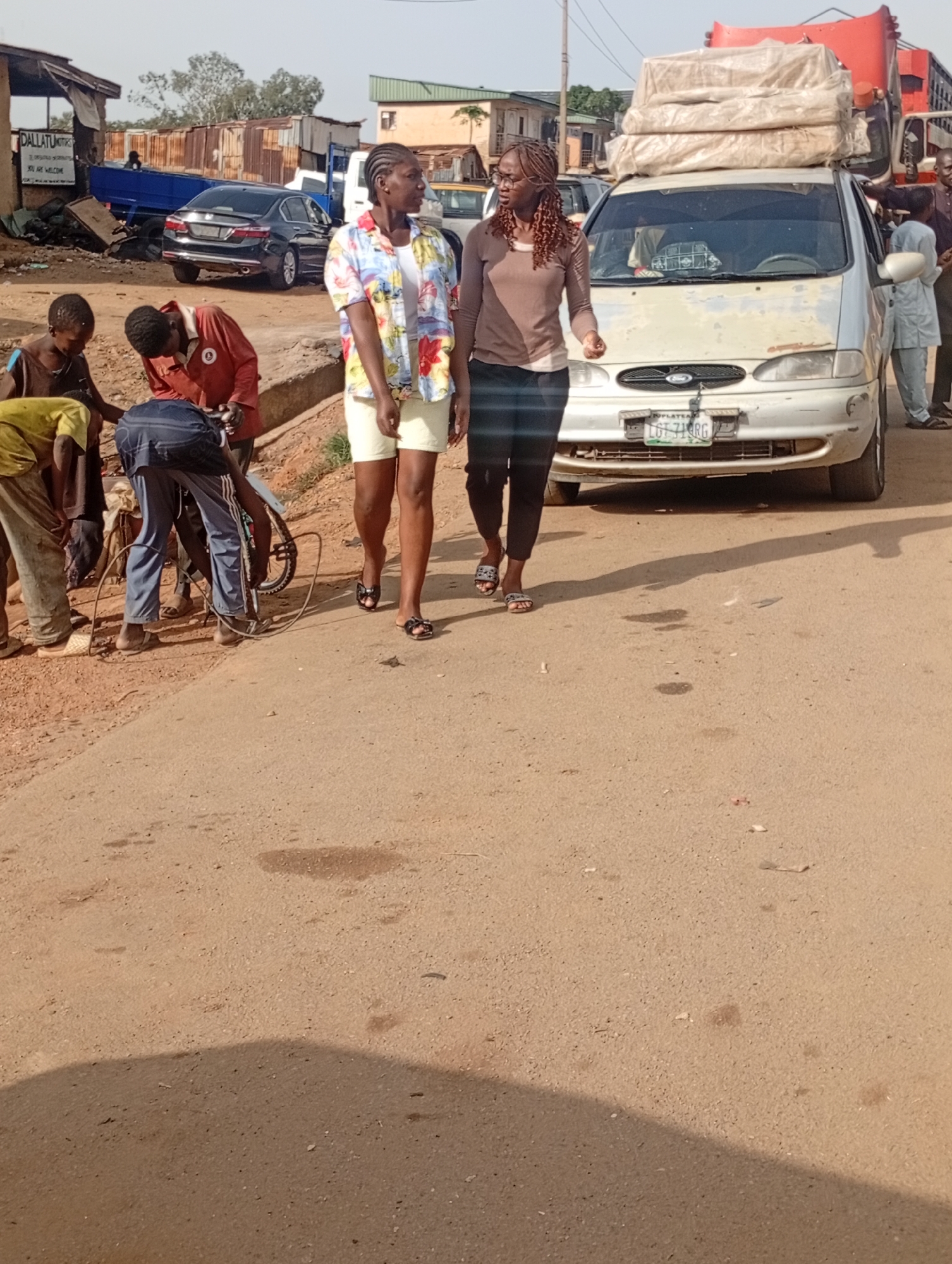 Two young women are walking on a dirt road in what appears to be a rural or semi-urban area in Rafin Par, Nigeria. The woman on the left wears a colorful floral short-sleeved shirt and light-colored shorts, while the woman on the right is dressed in a brown long-sleeved shirt and dark pants. Both are wearing sandals.
Behind them, several children are gathered around a bicycle, with one child in a red shirt appearing to work on it. Another child in a yellow shirt is also present. In the background, a white van is parked, loaded with what looks like mattresses or bedding strapped to its roof. A red truck is visible to the right of the van. Further in the background, there are buildings with corrugated metal roofs and some other vehicles.
The scene suggests a busy street or market area. The time of day appears to be daytime, likely morning or afternoon, given the sunlight and shadows. The weather seems warm and dry, indicated by the dusty road and the clothing of the people. The overall atmosphere seems casual and everyday.
There is some visible text on a sign in the far background to the left, which reads "DALLATU MOTORS". The license plate on the white van is partially visible and appears to read "LGT 711 RG".
