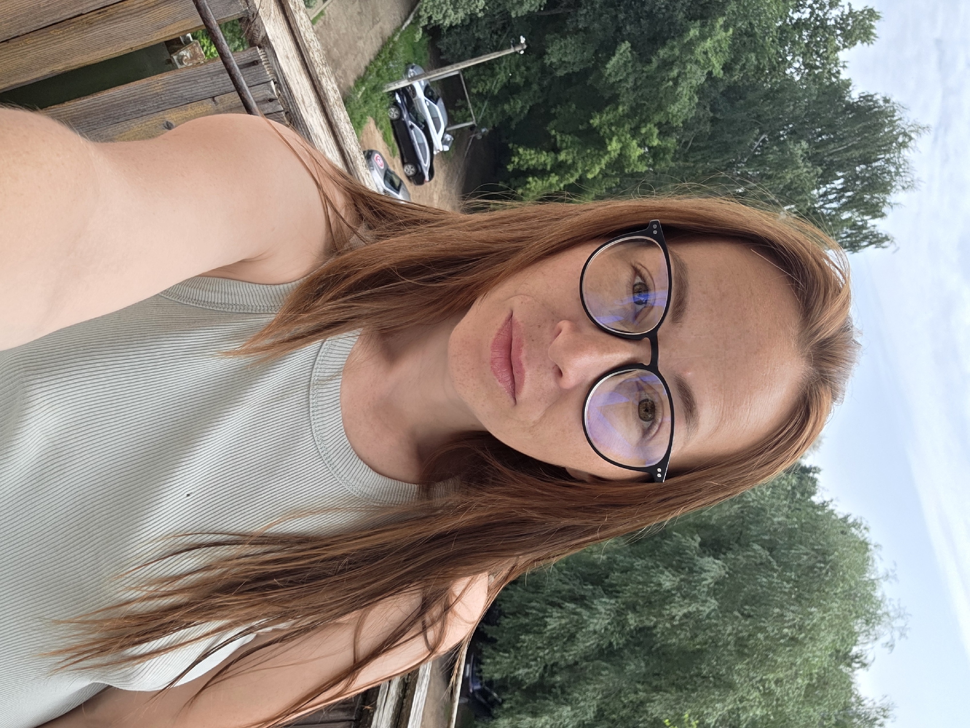 The image is a selfie taken outdoors. A woman with long auburn hair and glasses is the main subject, positioned centrally and facing the camera. She is wearing a light green ribbed tank top. In the background, there is lush green foliage from trees and a partly cloudy sky. Below the woman, a wooden railing and structure are visible, suggesting she is on a balcony or elevated platform. Further in the background, there are several parked cars and a pole with a light fixture. The scene appears to be in a natural or semi-rural setting within the city of Chaikovsky, Russia, possibly near a residential area given the presence of cars and a building structure.