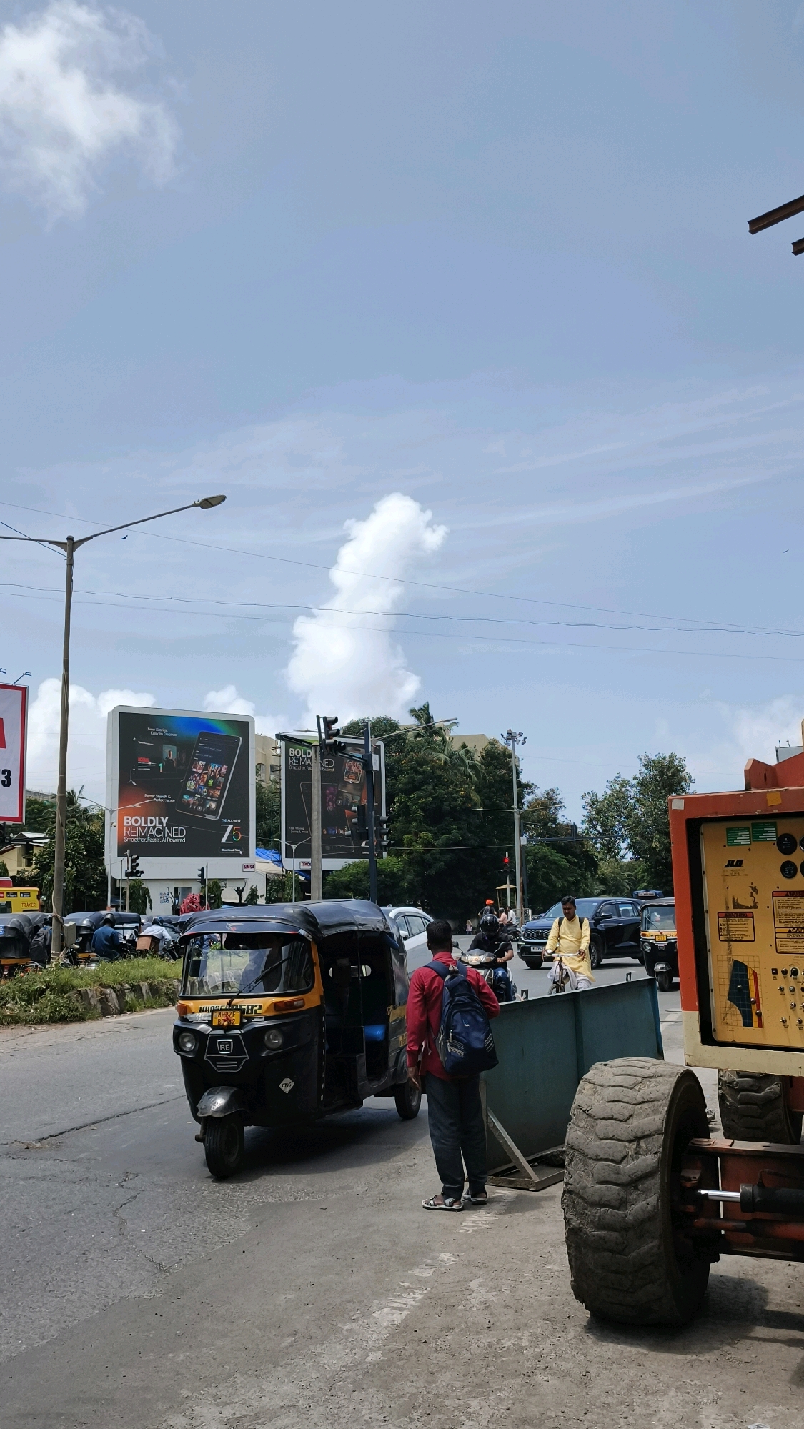 The image shows a street scene in Mumbai, India, under a partially cloudy sky. In the foreground, an auto-rickshaw is parked on the left side of the road, facing away from the camera. A man wearing a red shirt and a blue backpack stands to the right of the auto-rickshaw, facing it. He appears to be looking at something within the auto-rickshaw. Further ahead on the road, several vehicles, including cars and motorcycles, are visible, some in motion and some stopped, suggesting traffic.
On the right side of the image, a large piece of heavy machinery with a substantial tire is parked. Beyond the road, billboards advertising "BOLDLY REIMAGINED Z5" are prominently displayed. The surrounding environment includes trees and buildings, indicating an urban setting. The overall impression is of a busy street corner in Mumbai, with a mix of pedestrian and vehicular activity, set against the backdrop of commercial advertising.